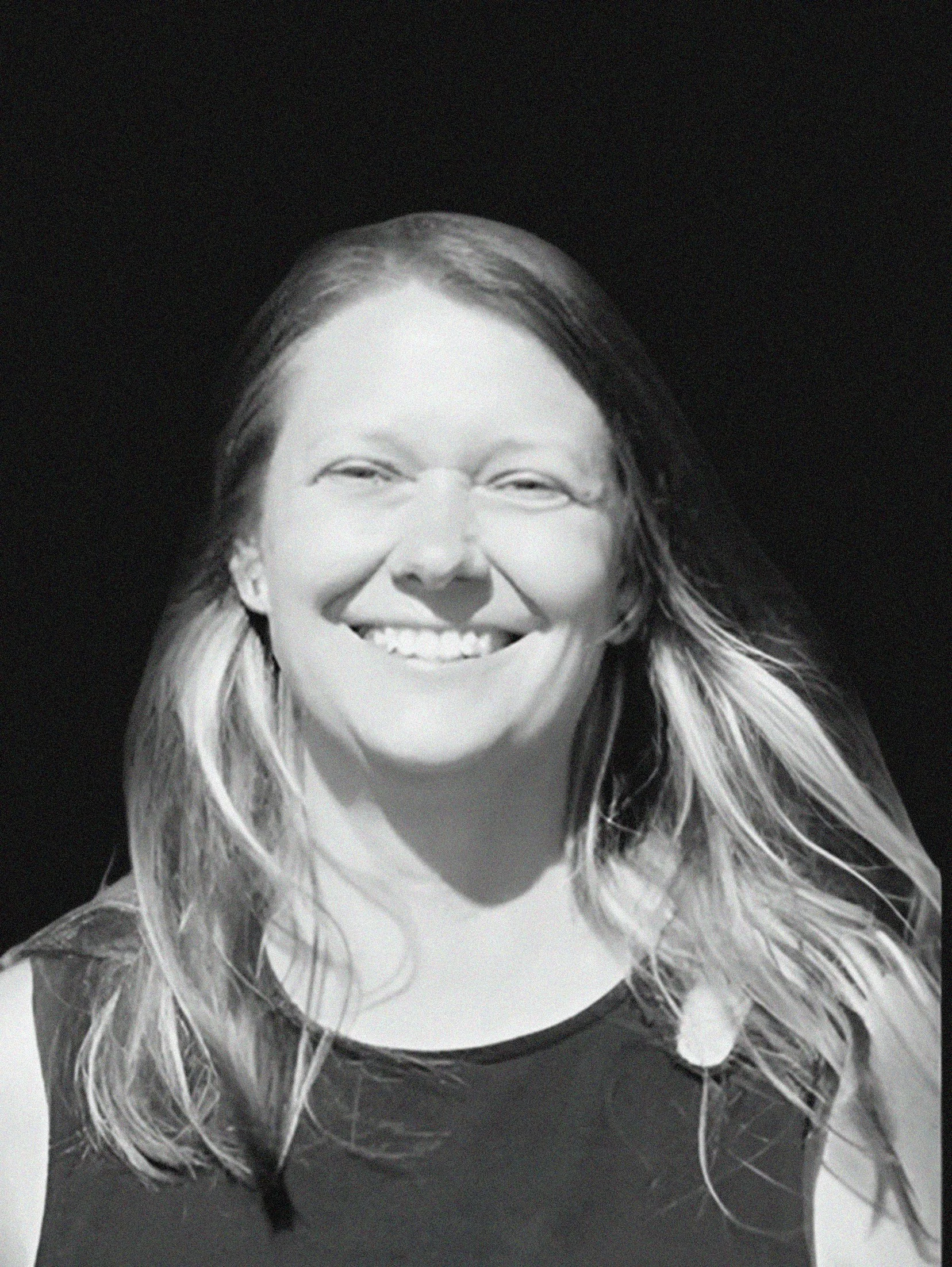 Black and white photo of a smiling woman with long hair, standing outdoors with a building and trees in the background.