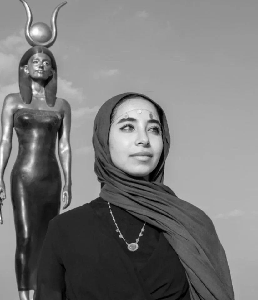 A woman wearing a headscarf and jewelry standing outdoors with a tall statue of an Egyptian goddess with a headdress featuring a sun disk and horns in the background.