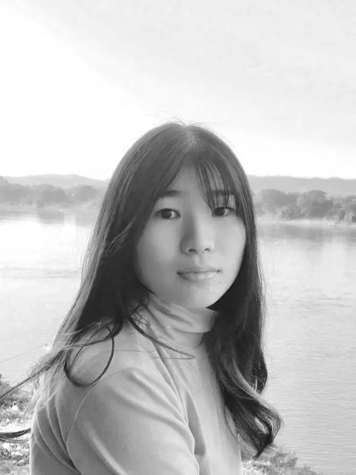 Black and white photo of a young woman with long dark hair, wearing a turtleneck, standing near a body of water with trees in the background.