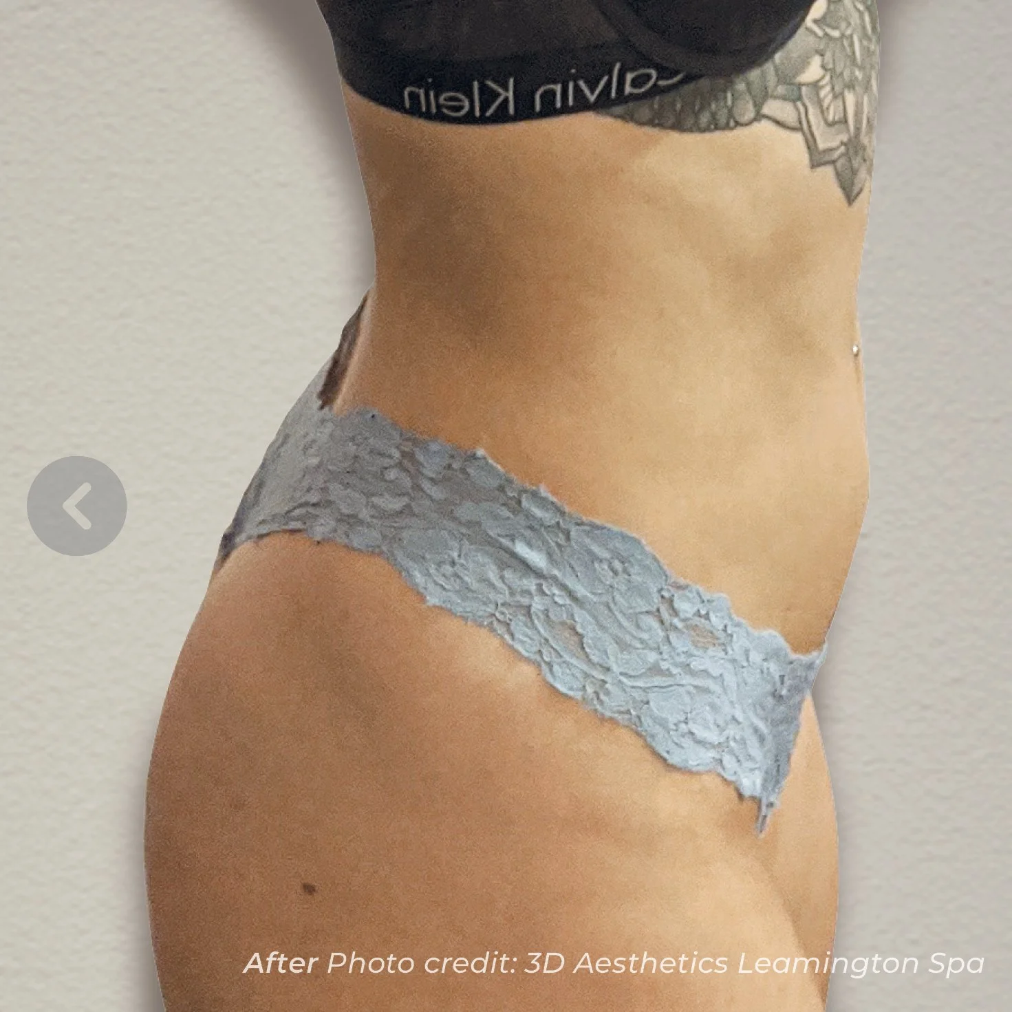 Close-up of a woman's lower abdomen and upper thigh, showing a line of grey textured body paint or tattoo resembling a torn piece of fabric on her hip and waist. She has a tattoo on her ribcage and a navel piercing, with the background being a plain