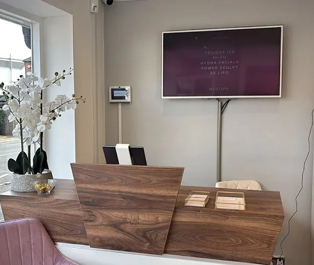 Reception desk with a wooden finish, a bouquet of white orchids on the left, a monitor on the wall displaying treatment information, and a window in the background.