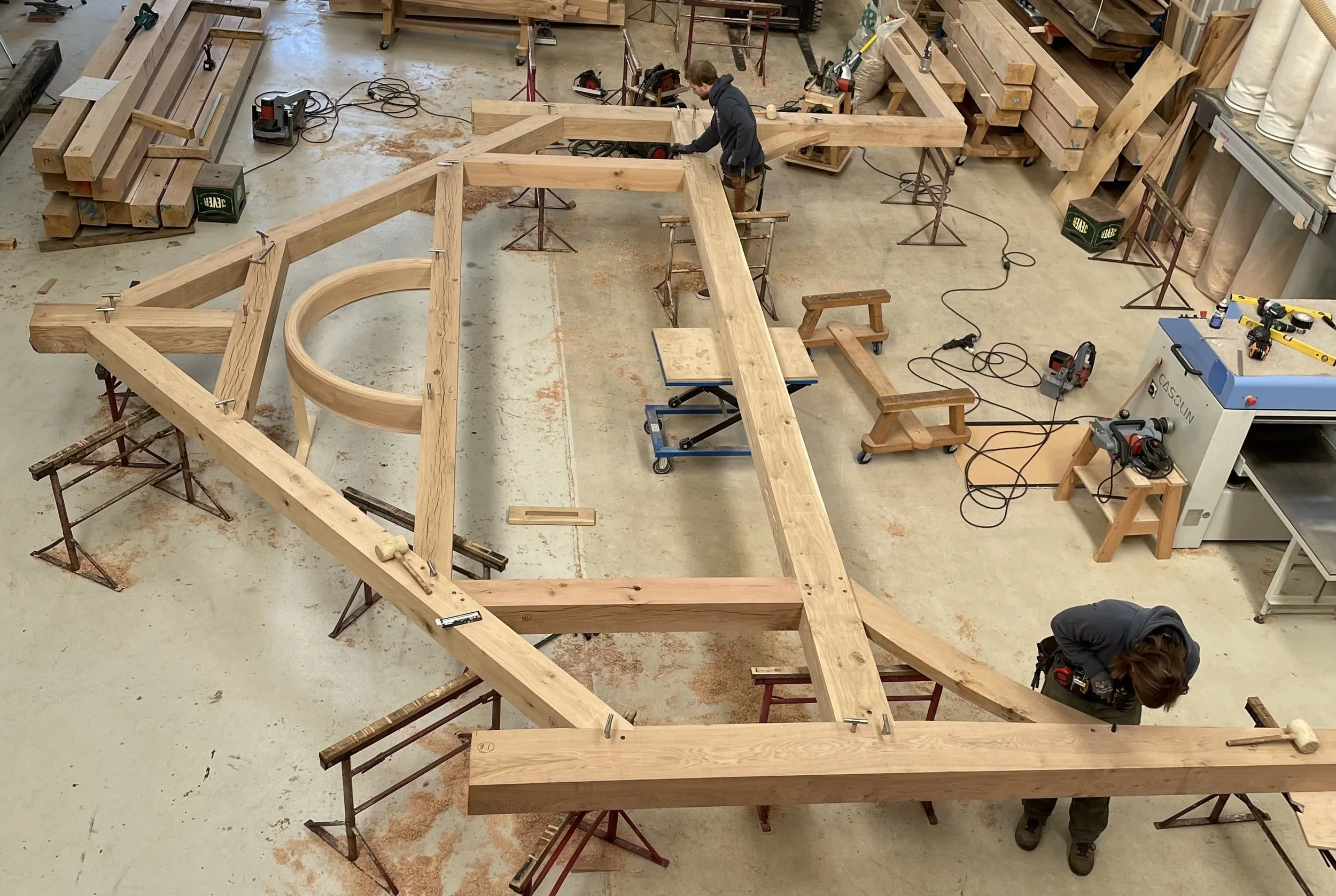 Two workers assembling a large wooden structure in a woodworking workshop. The structure includes circular and angular wooden pieces, supported on multiple workbenches and saw horses, with woodworking tools and machinery visible around them.