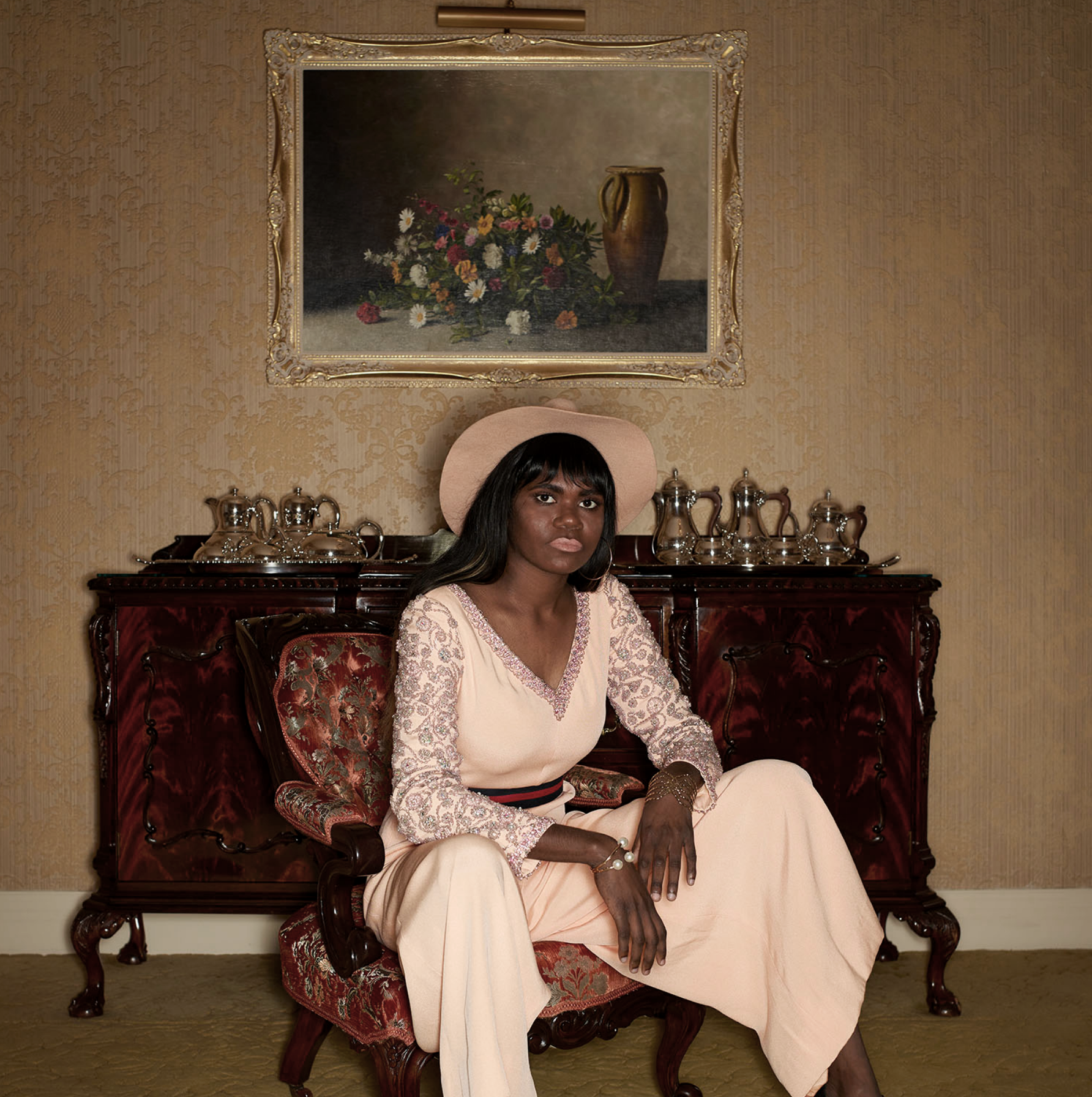 A woman with dark skin and long dark hair sits on an ornate wooden chair in a room with vintage decor. She is wearing a light pink wide-brimmed hat, a long-sleeved pink lace top, and a long light pink skirt. Behind her is a dark wooden sideboard with silver tea/coffee serving ware. A framed still-life painting of a vase and flowers hangs on the wall above her, which has gold patterned wallpaper.