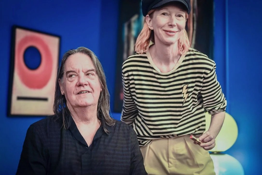 Two people, a woman sitting and a woman standing, inside a colorful room with blue walls and abstract artwork.