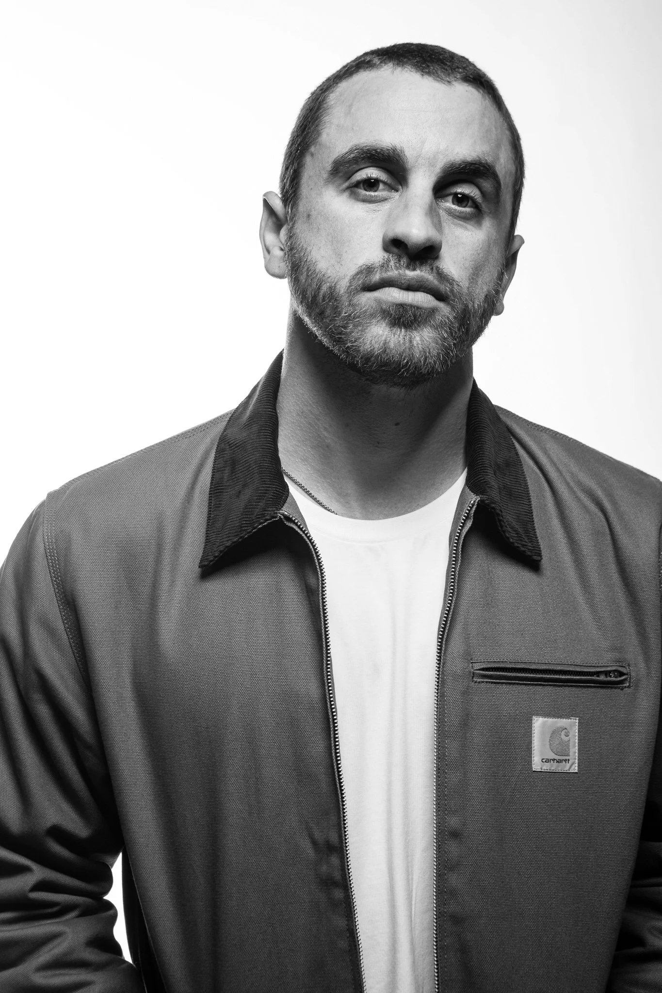 Black and white portrait of a man with short hair and a beard, wearing a zippered Carhartt jacket over a t-shirt, standing against a plain white background.