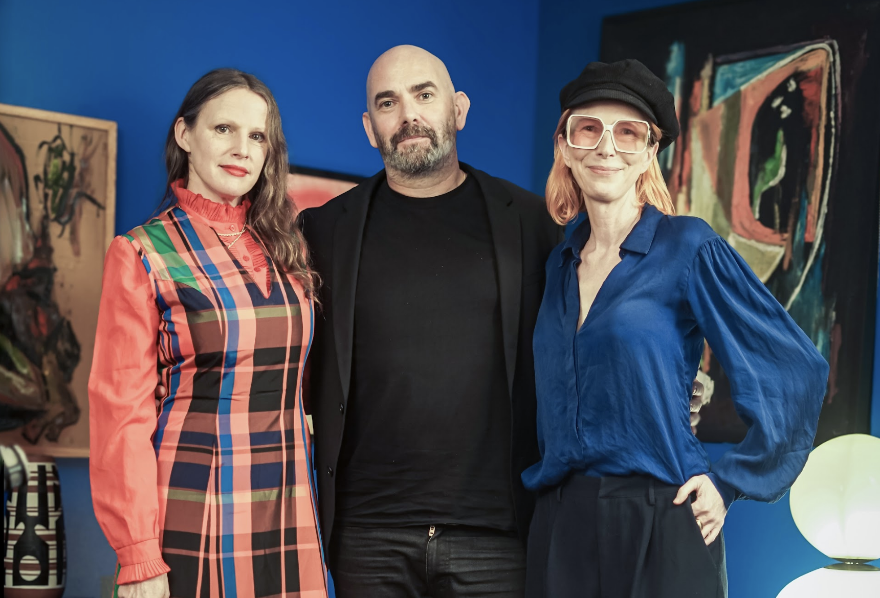 Three people standing together indoors with colorful paintings on the wall behind them.