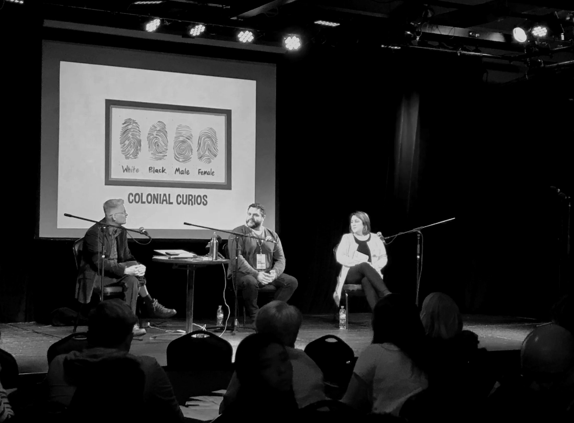 A panel discussion on stage with three people seated and microphones. A large screen behind them displays images of fingerprints labeled 'White', 'Black', 'Male', and 'Female', with the text 'Colonial Curious' beneath.