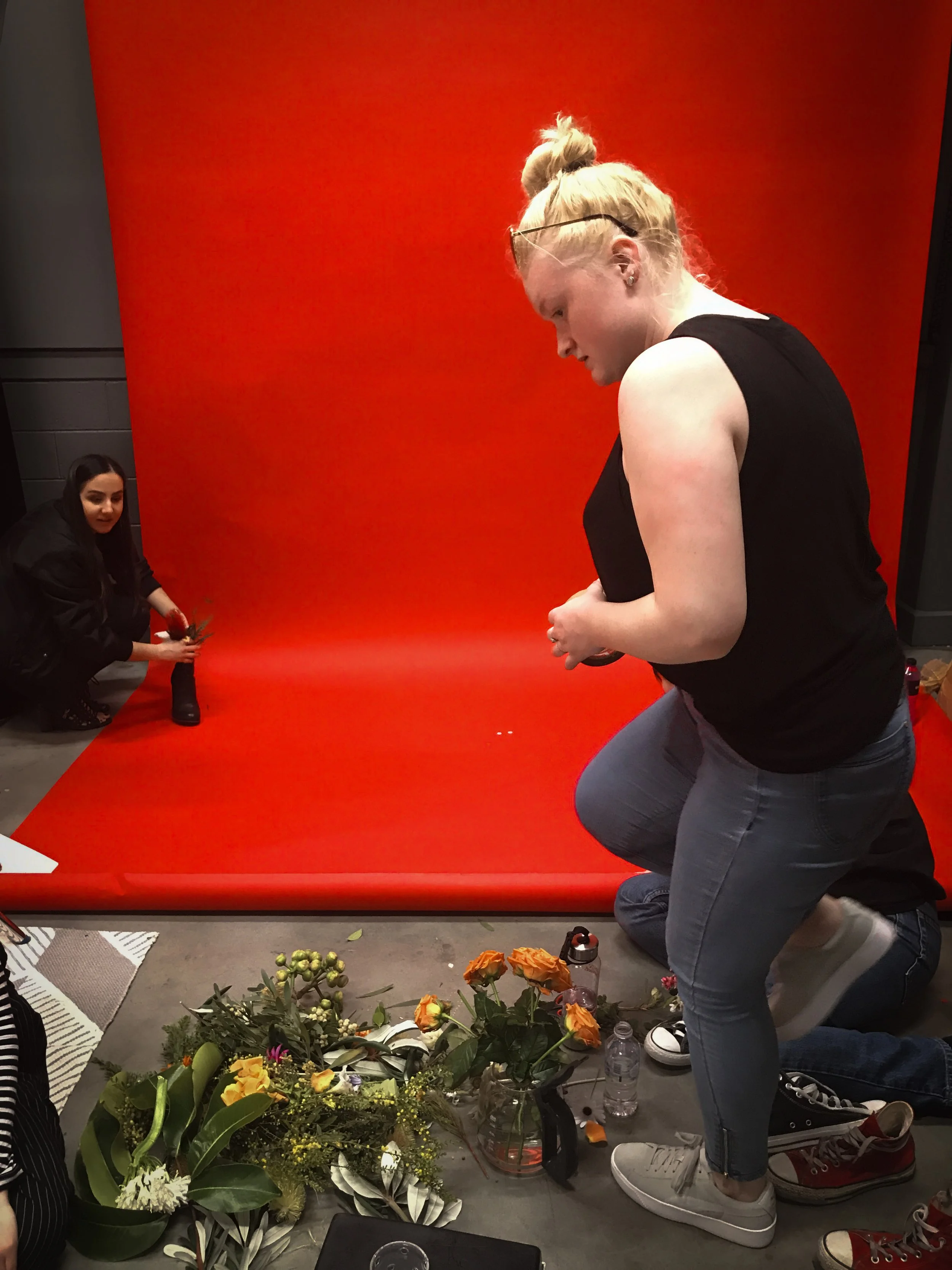 TRIPLE AXEL MEDIA LITERACY flowers, green leaves, and ferns in shoes with the text 'The Language of FlTwo women preparing for a photo shoot, one kneeling and the other standing in front of a red backdrop, with flowers and water bottles on the ground.