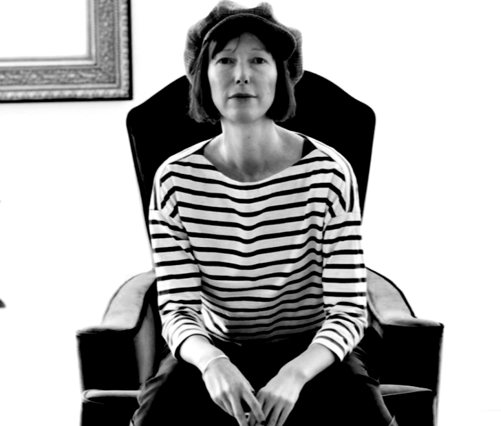 A woman with short hair wearing a striped shirt and a beret, sitting in a high-backed armchair in a room with a framed picture on the wall.