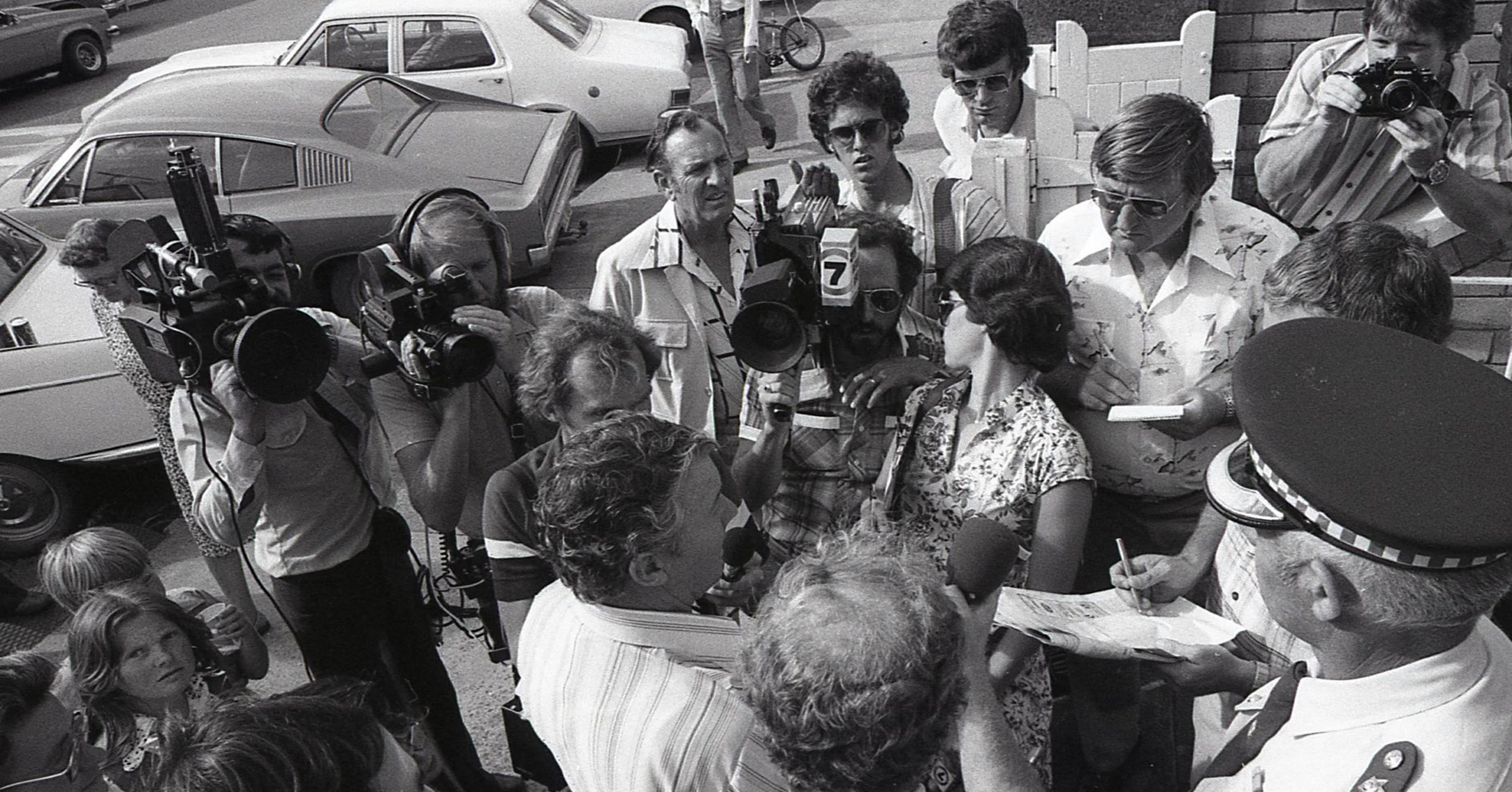 Group of journalists and photographers surrounded a woman as she speaks into microphones outside in a parking lot with vintage cars in the background.