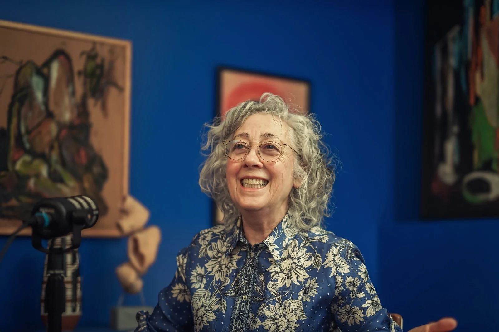 A woman with curly gray hair and glasses smiling in front of a blue wall with paintings.