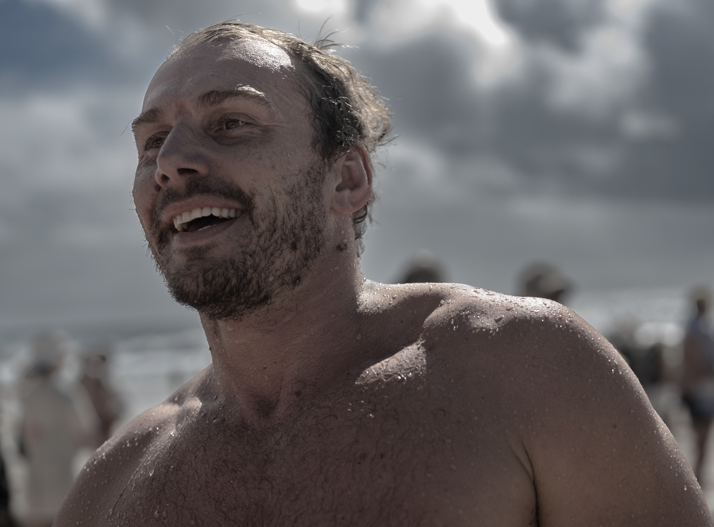 Close-up of a smiling man with wet hair and water droplets on his skin, outdoors on a cloudy day.