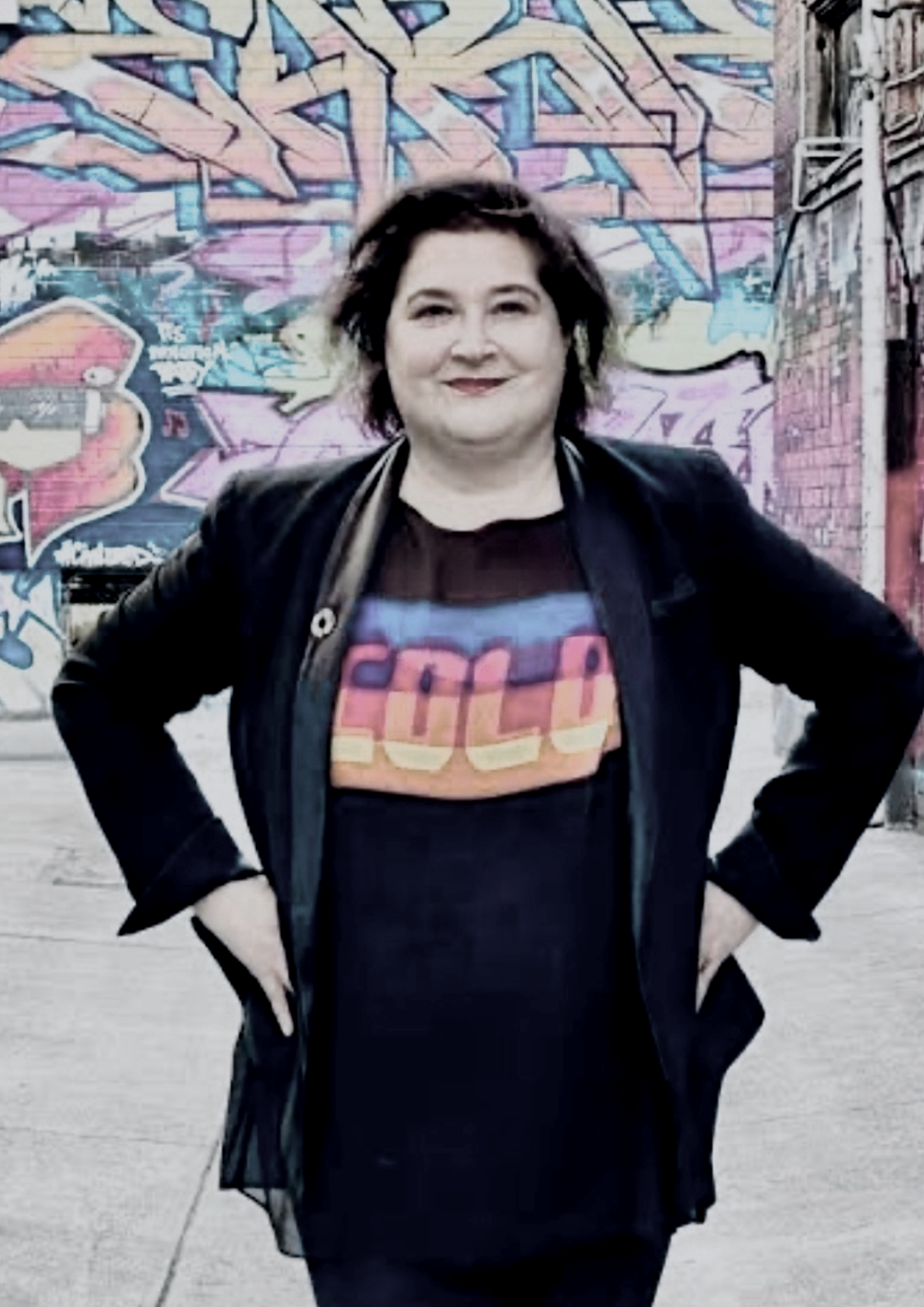 Woman standing in front of a colorful graffiti wall, wearing a black jacket and a shirt with a rainbow-colored logo.