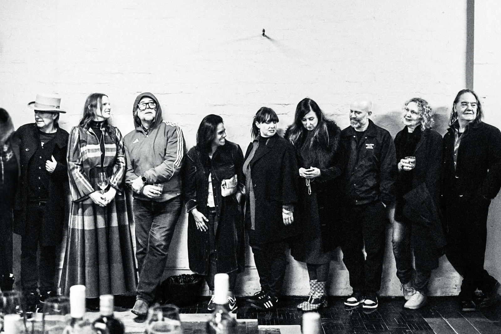 Group of nine people standing against a wall at a social gathering, some holding drinks, chatting and smiling.