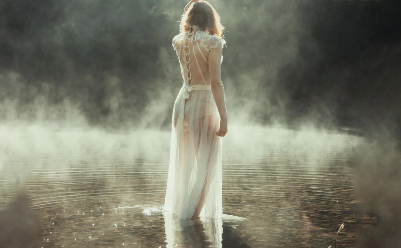 A woman in a white, semi-transparent dress standing in a foggy lake, with her back to the camera and sunlight illuminating her from behind.