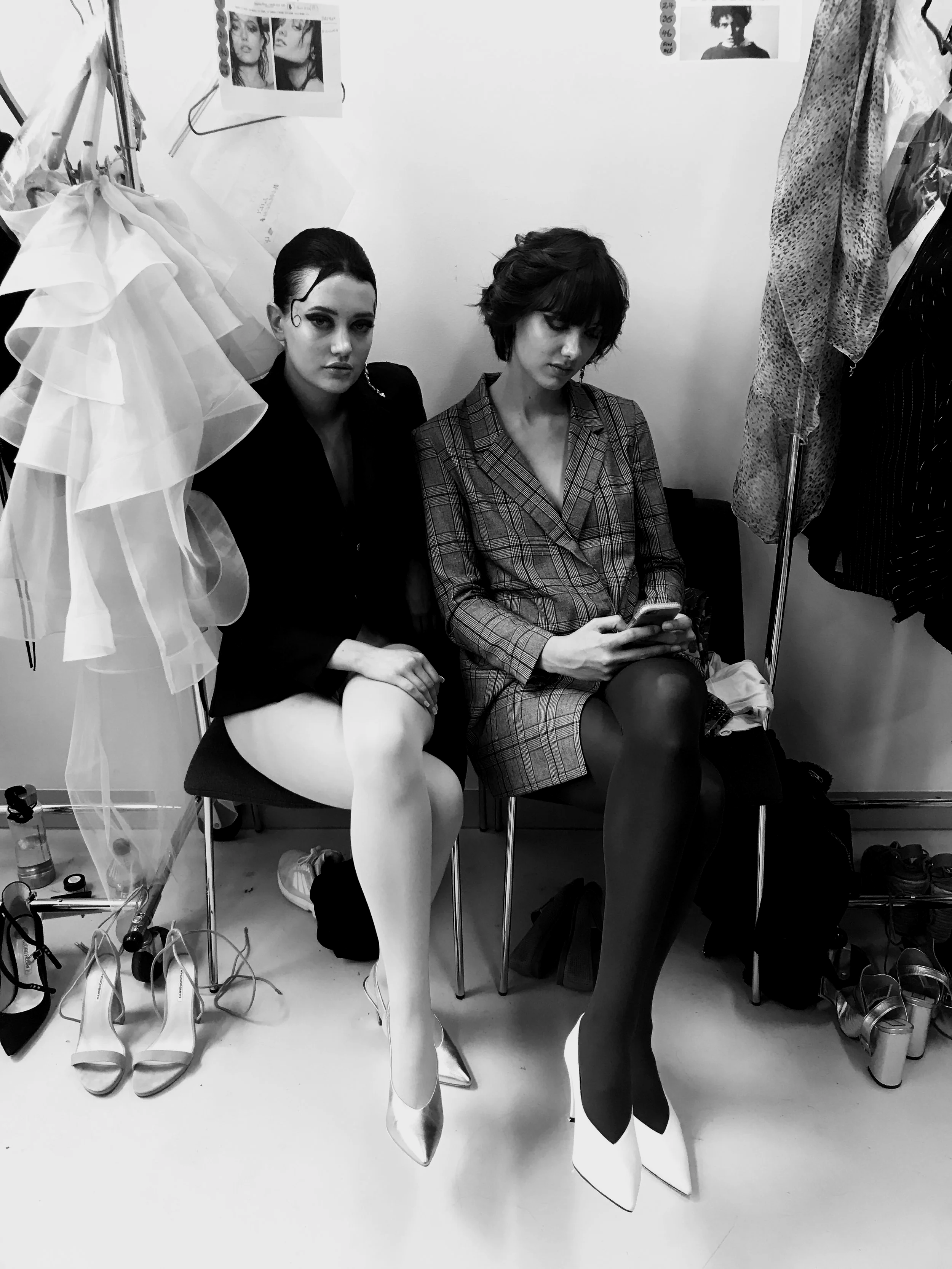 Two women sitting on chairs backstage, one looking at her phone. The woman on the left has dark hair, a dark blazer, and white tights. The woman on the right has short hair, a checked blazer, and black tights. Shoes and clothing items are visible around them.