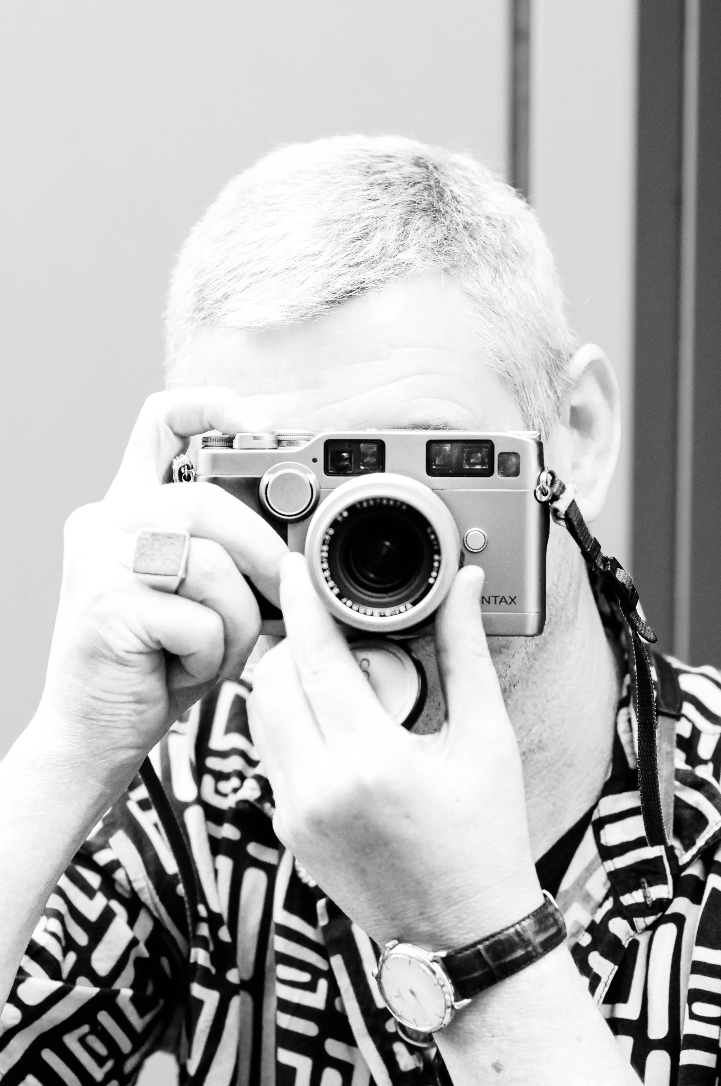 A person with short, gray hair taking a photo with a camera, wearing a patterned shirt and a wristwatch.