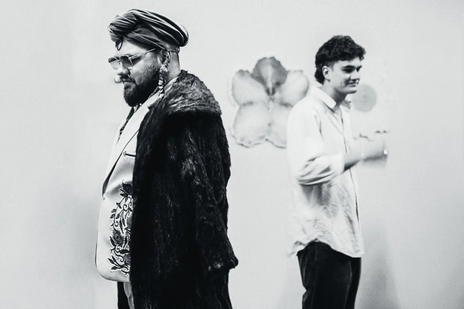Black and white photo of two men standing apart against a plain wall with floral artwork. The man on the left has a beard, sunglasses, ornate earrings, a turban, and wears a fur coat with a decorated bag over his shoulder. The man on the right has curly hair, wears a light-colored shirt, and appears to be smiling.