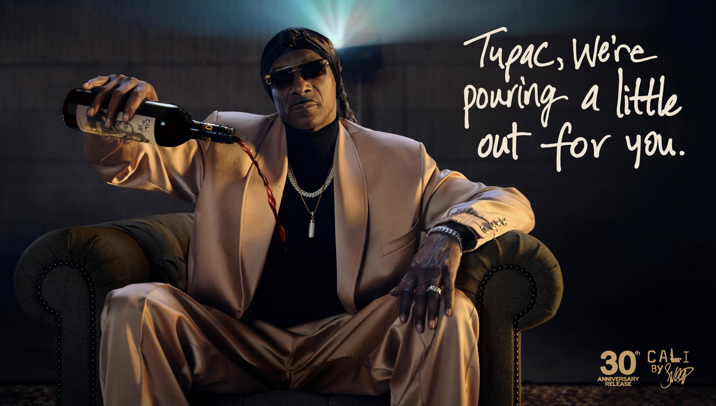 A man dressed in a beige suit, black turtleneck, wearing sunglasses, jewelry, and a head covering, sitting on a green armchair, pouring red wine from a bottle with a label reading 'Cal G.