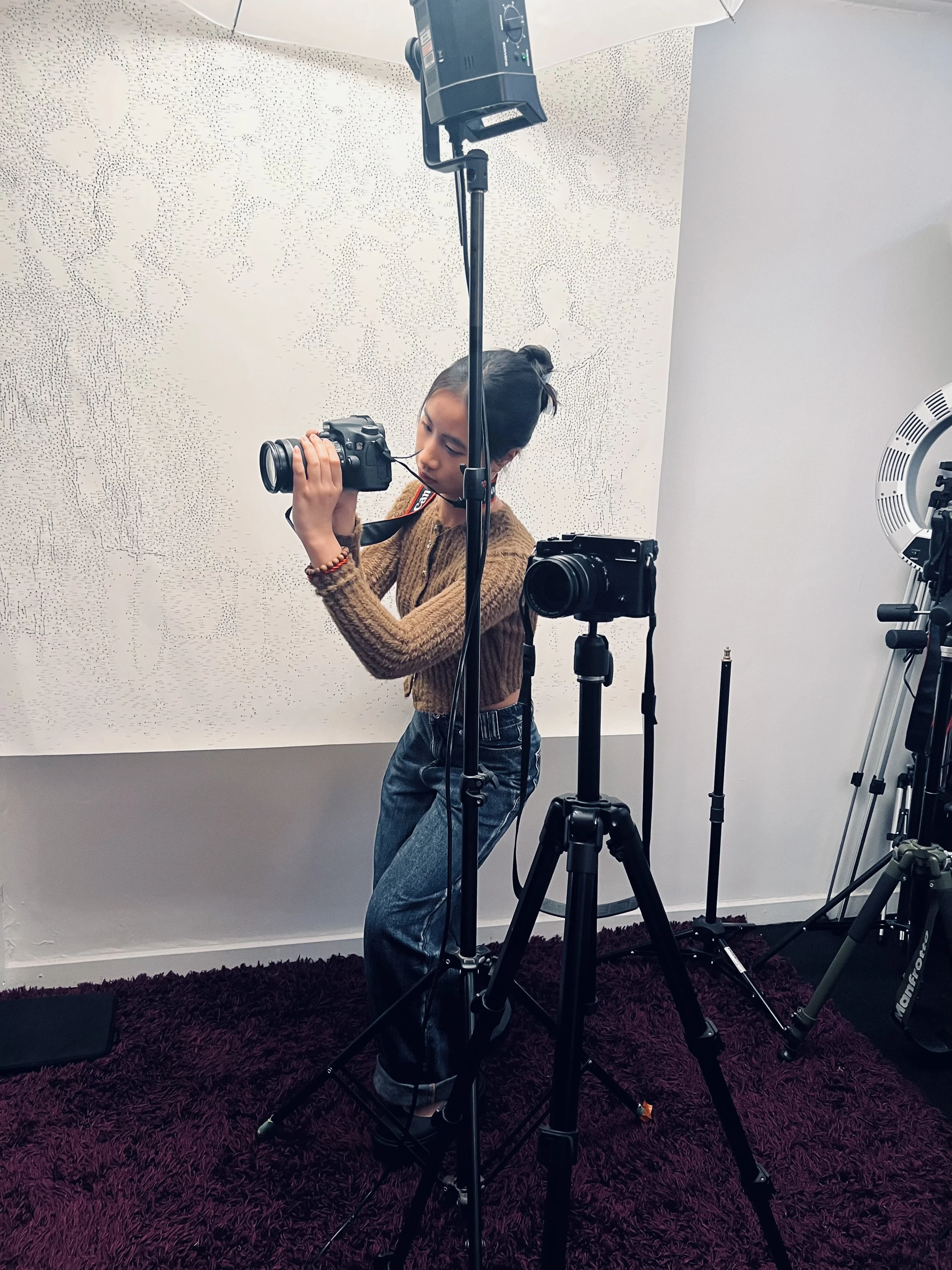 A young woman with a bun hairstyle, wearing a brown sweater and jeans, is testing a camera on a tripod in a photography studio with lighting equipment and a textured white backdrop.