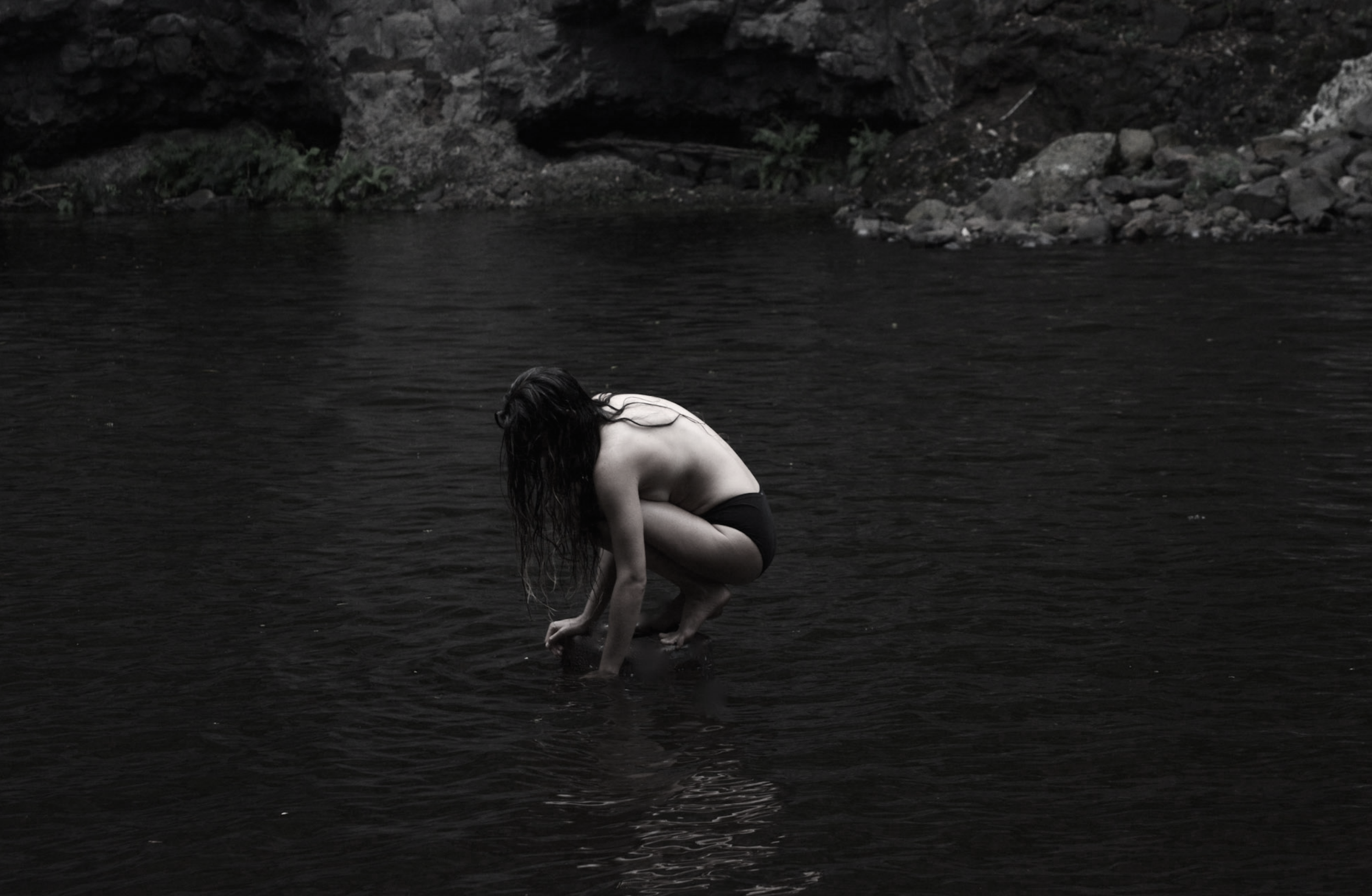 A person with long dark hair crouching in a body of water, with rocks and a rocky shoreline in the background.