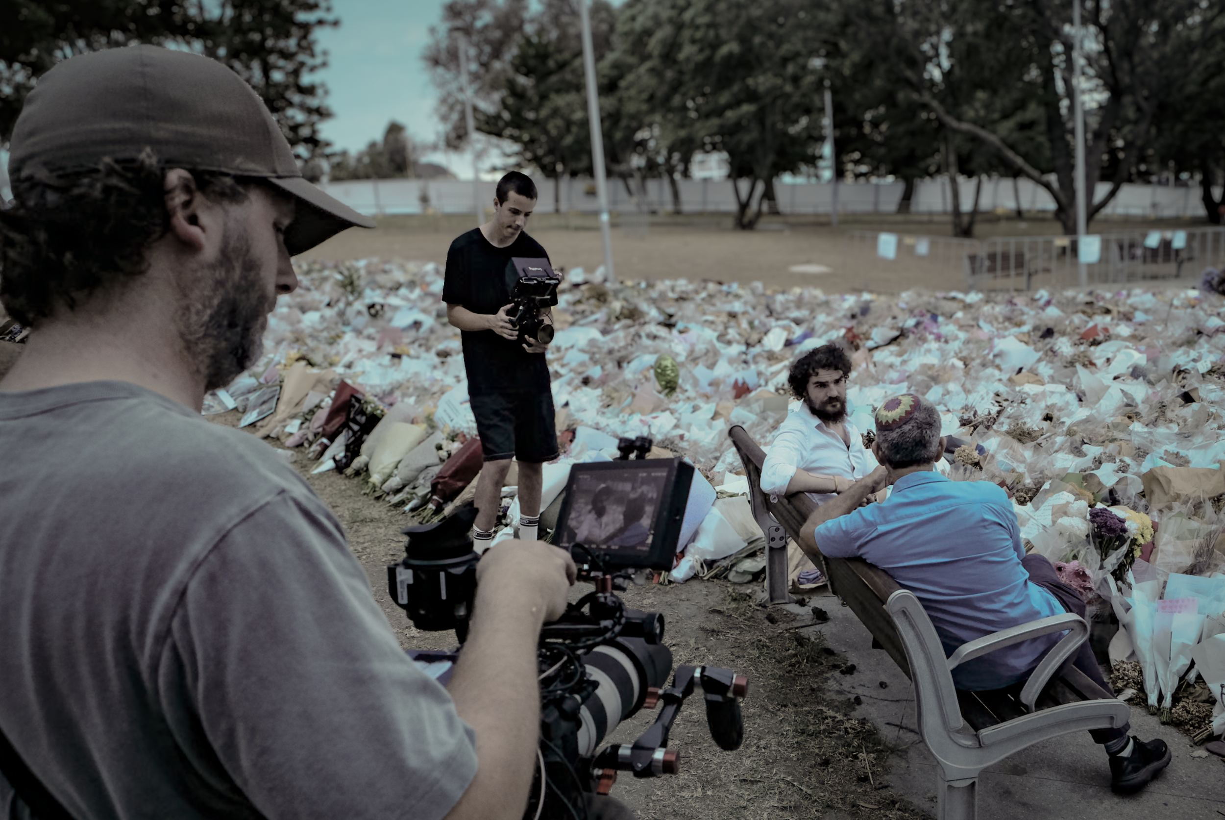 Filming crew recording in front of memorial with flowers and messages at a park.