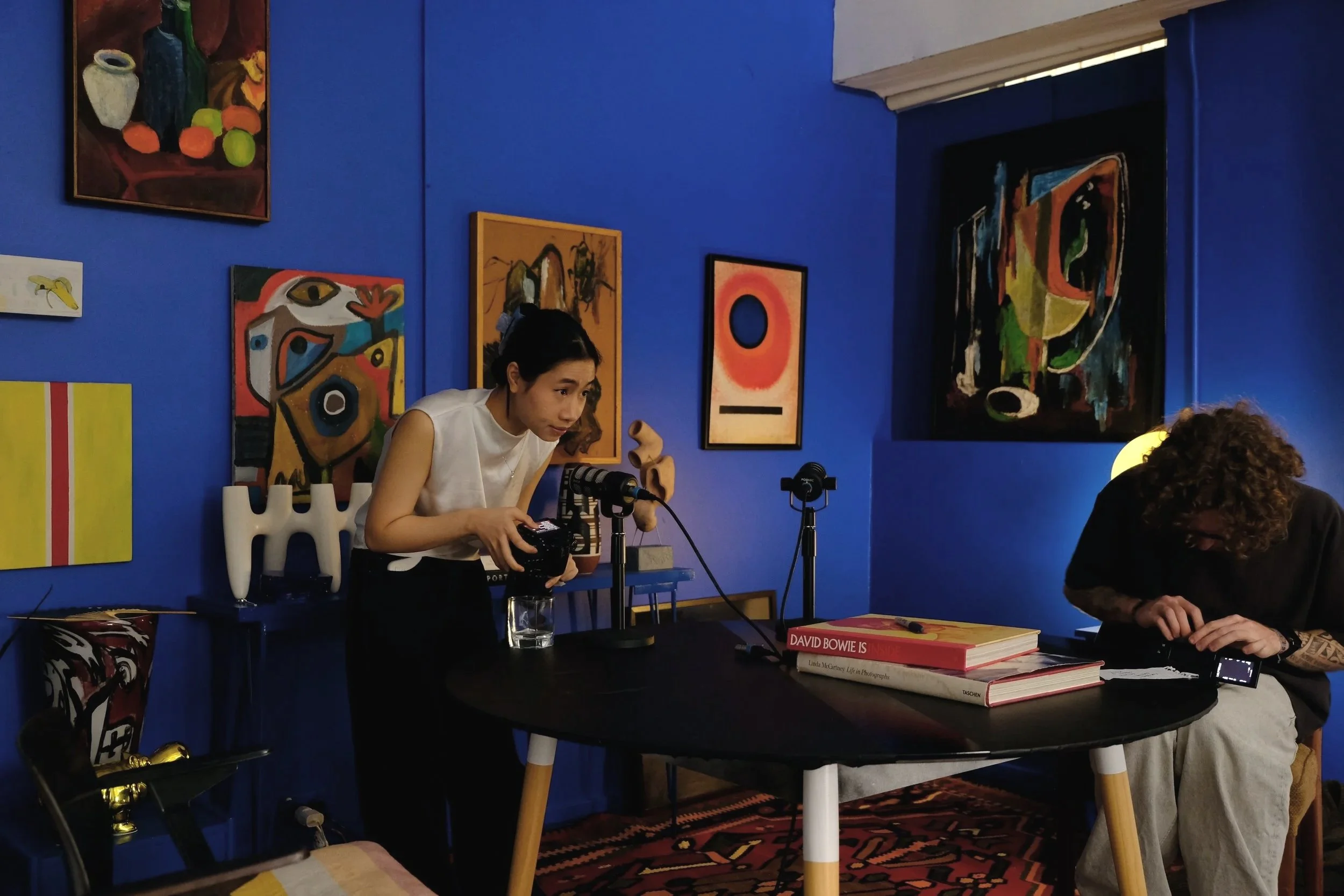 A mentor woman adjusting recording equipment at a table in a room with blue walls decorated with colorful abstract paintings; another person sits at the table, looking down, with books and equipment on the table.