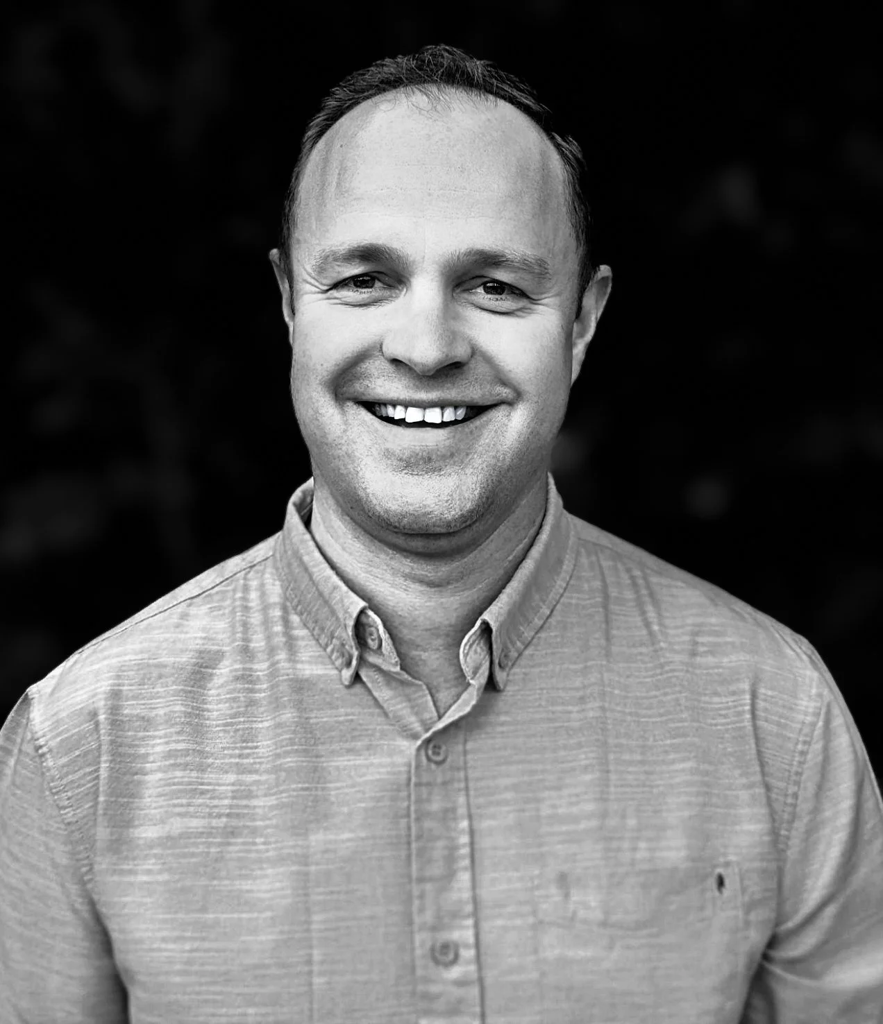 A smiling man with short hair and a collared shirt, posing against a dark background.