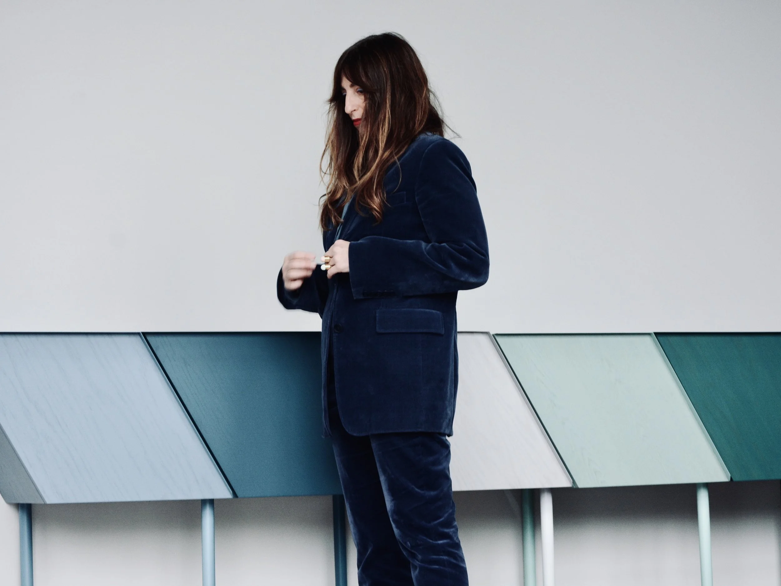 A woman with long brown hair, wearing a dark blue velvet blazer and pants, standing against a gray wall and looking down while adjusting her blazer.