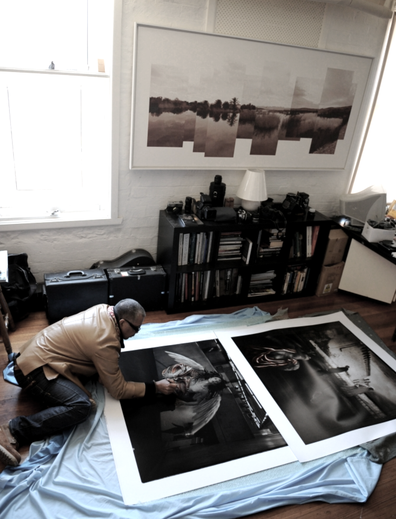 A man kneels on the floor, working on two large framed photographs of a black and white angel sculpture and an industrial scene, with a black bookshelf filled with books and cameras behind him.