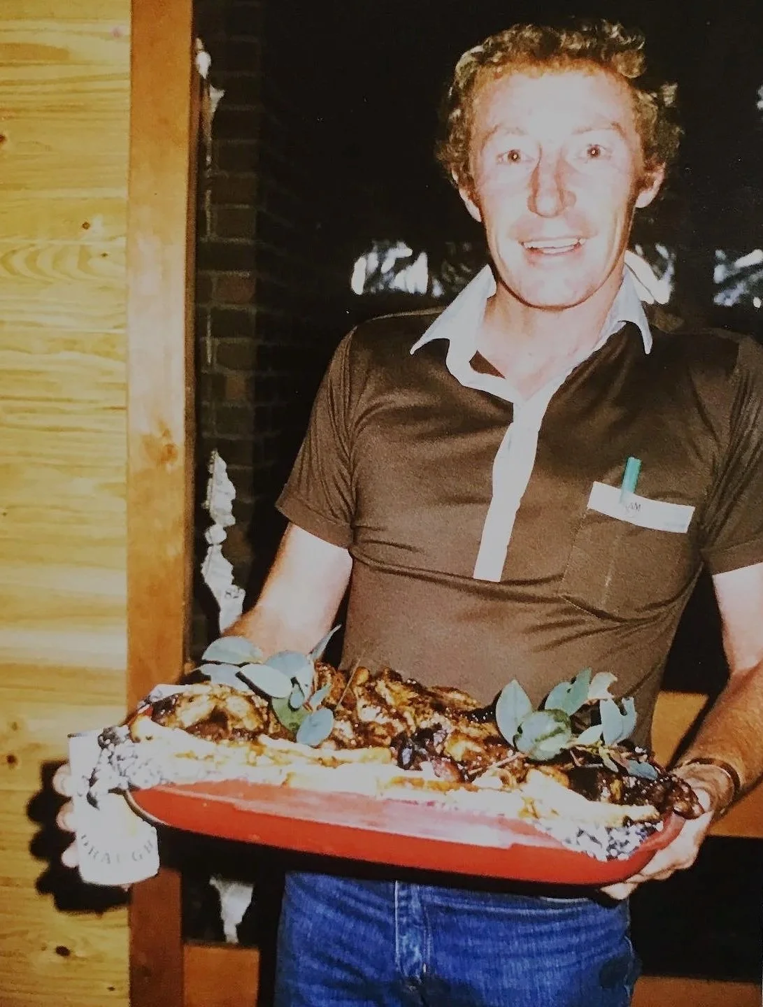 A man holding a large dish of food, possibly pizza or lasagna, garnished with leaves, indoors with wooden and brick background.