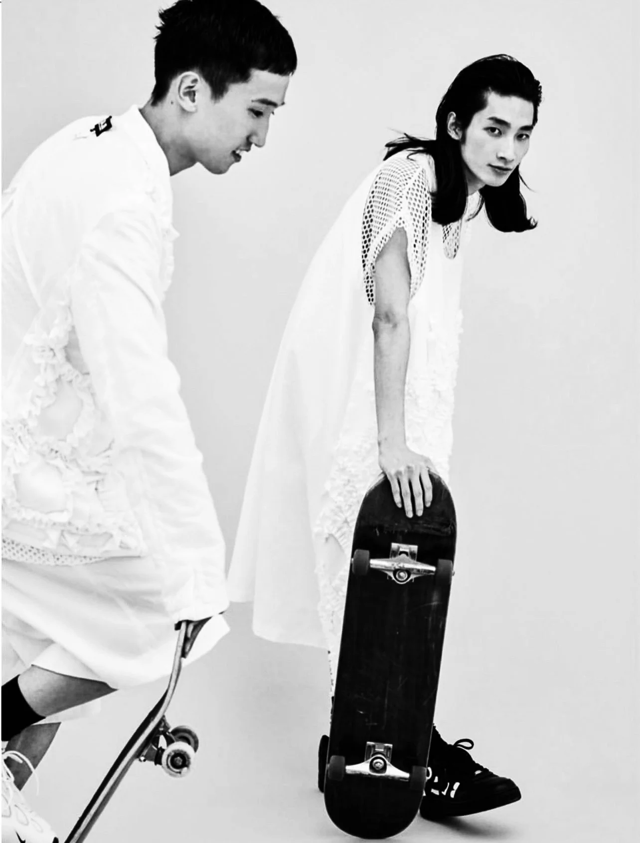 Two people dressed in white, one male and one female, are leaning forward. The female is holding a skateboard and looking at the camera, while the male is on a skateboard and looking downward. The image is in black and white.