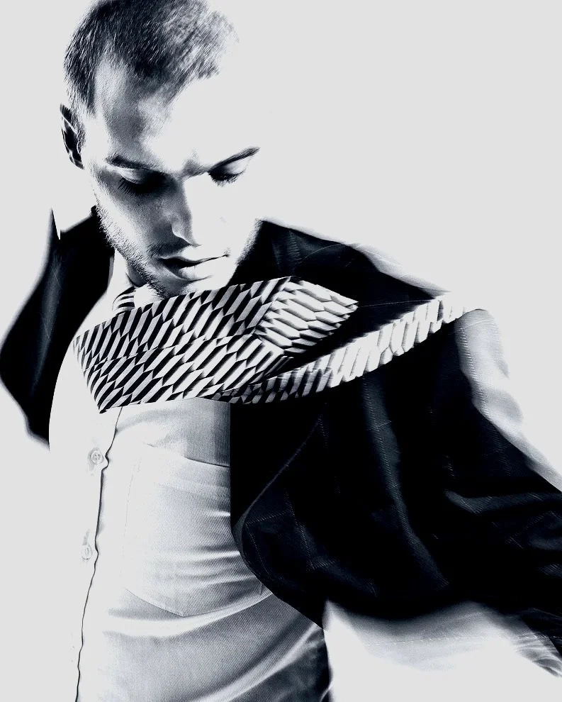 Black and white photo of a man looking down, wearing a patterned scarf, a button-up shirt, and a jacket.