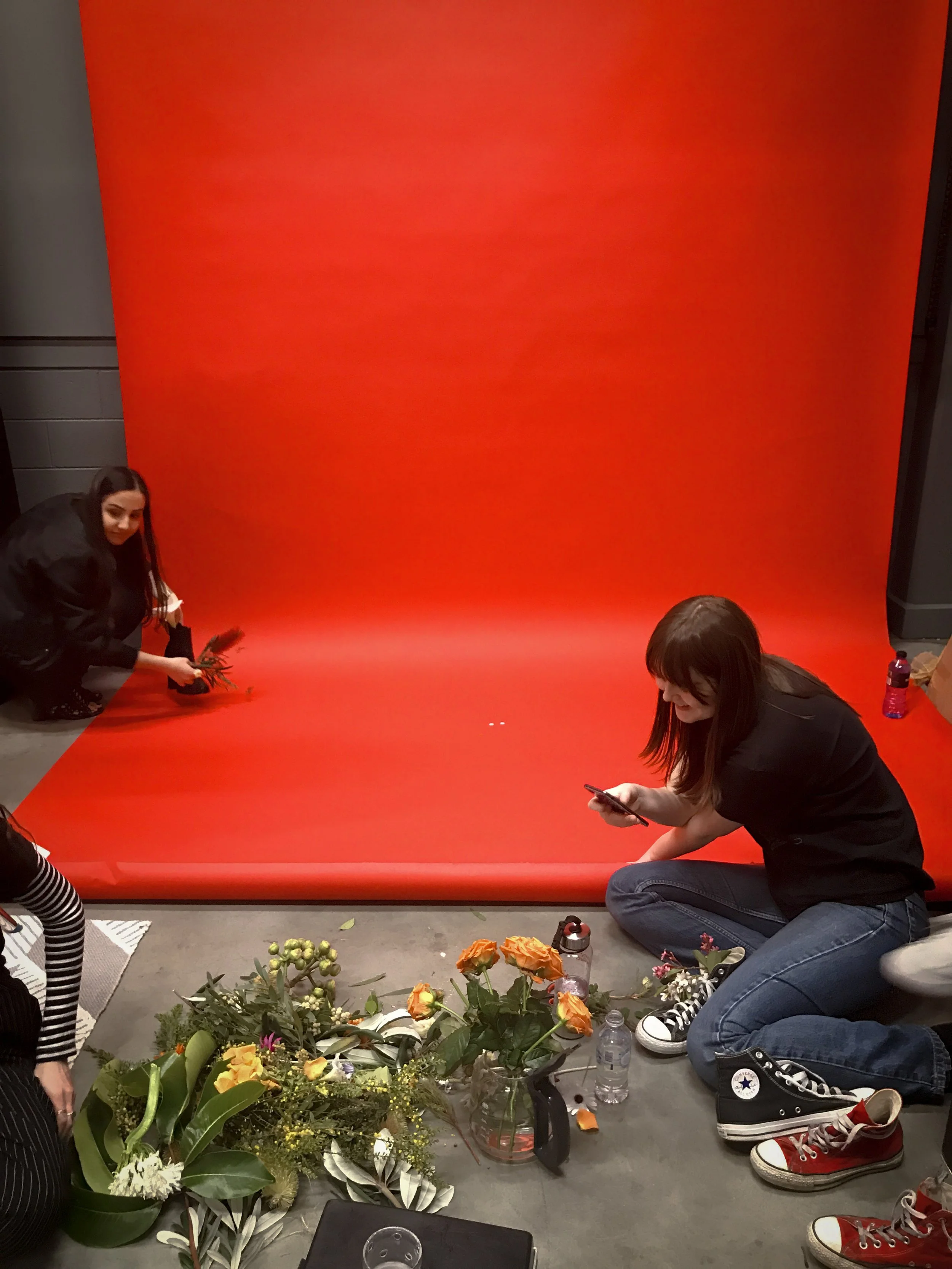 TRIPLE AXEL MEDIA LITERACY flowers, green leaves, and ferns in shoes witTwo women sitting on the floor in front of a large red backdrop, surrounded by flowers, water bottles, and footwear. One woman is holding a phone, the other is arranging flowers.