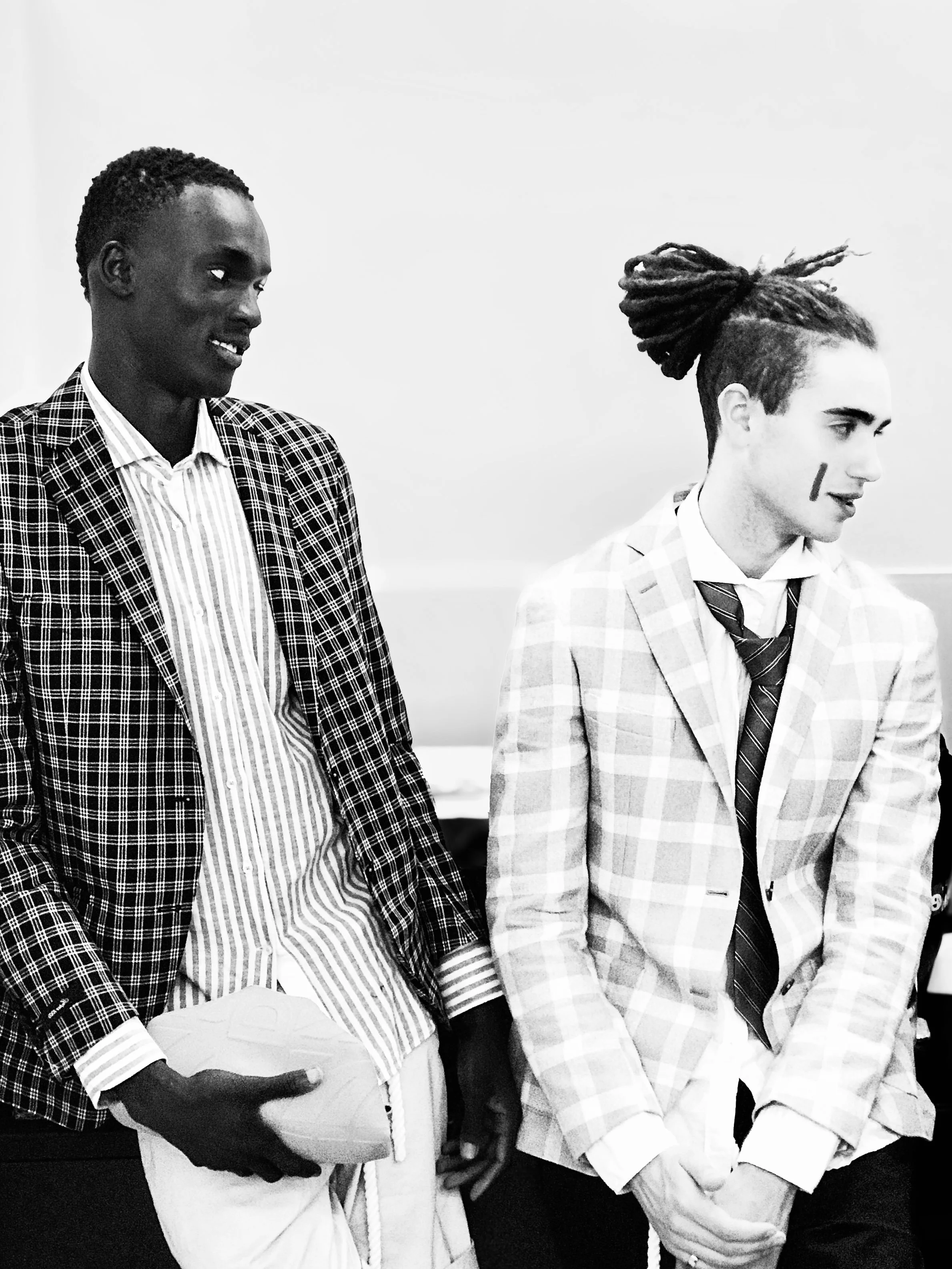 Two men in suits standing and talking, one holding a football, in a room with plain walls.