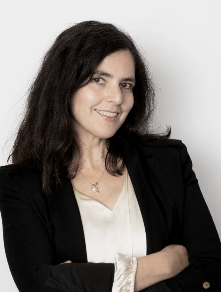 A woman with shoulder-length dark brown hair, smiling, wearing a black blazer over a cream blouse, with a pearl necklace, standing against a white background.