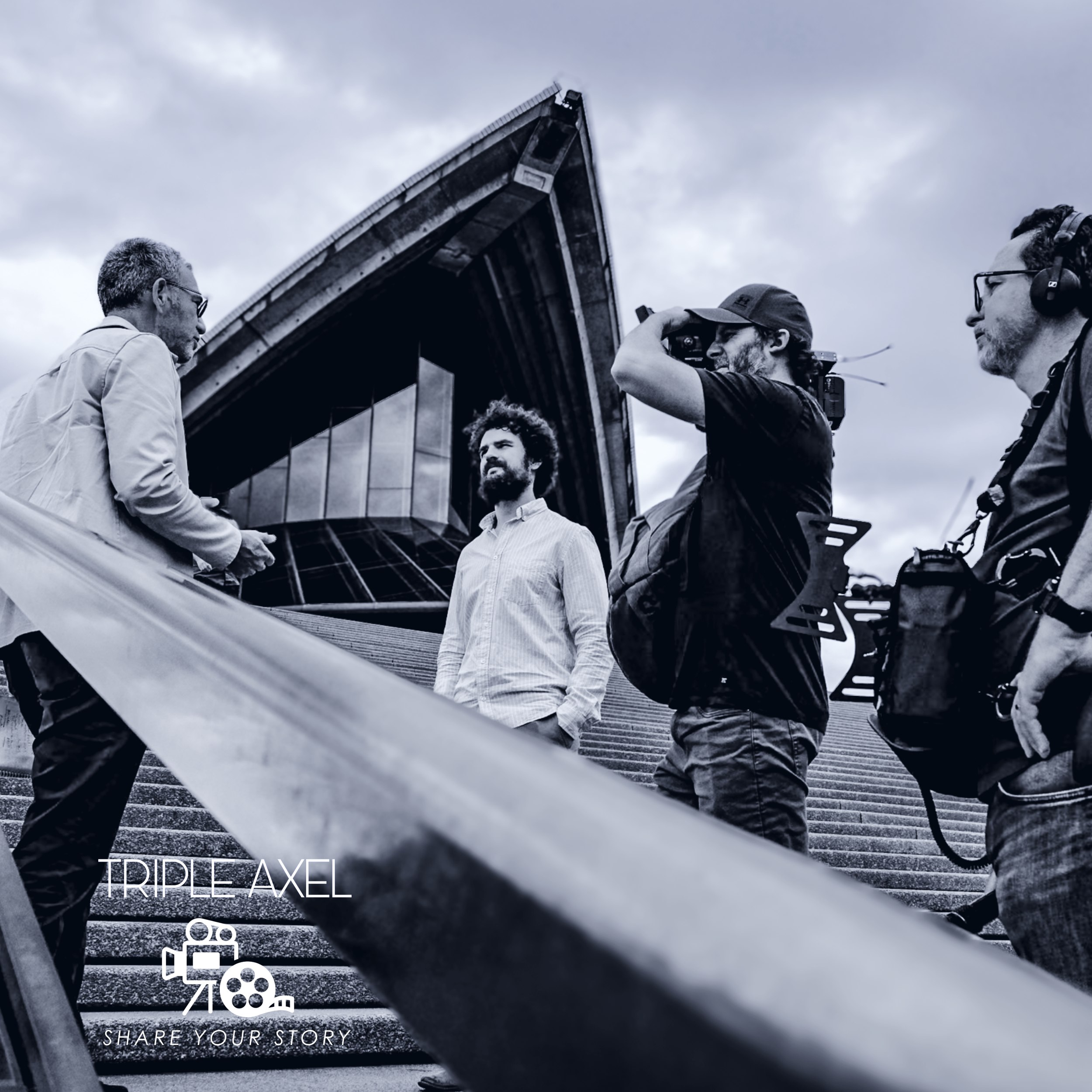 People standing on stairs behind a metal railing, some wearing headphones, with a large structure and cloudy sky in the background.