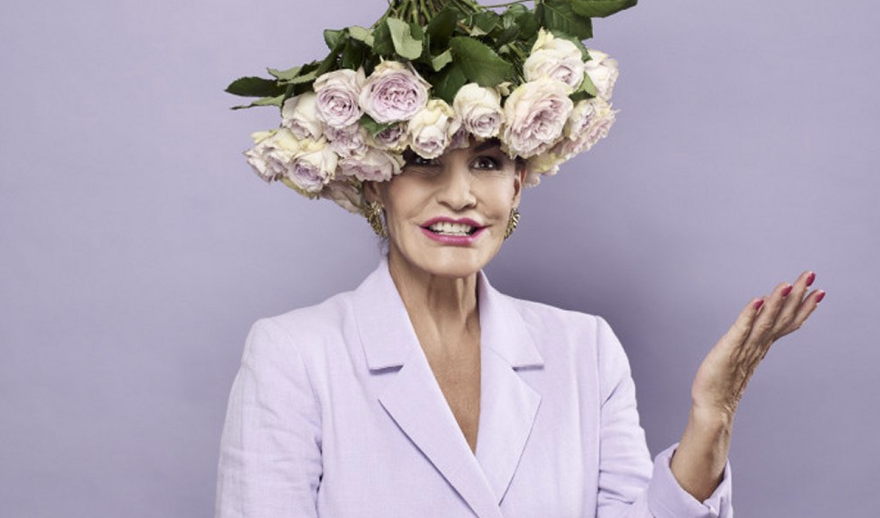 An creative industry leader, Mentor woman with a large hat made of pink roses, wearing a light purple blazer, smiling with pink lipstick, and gesturing with her hand.