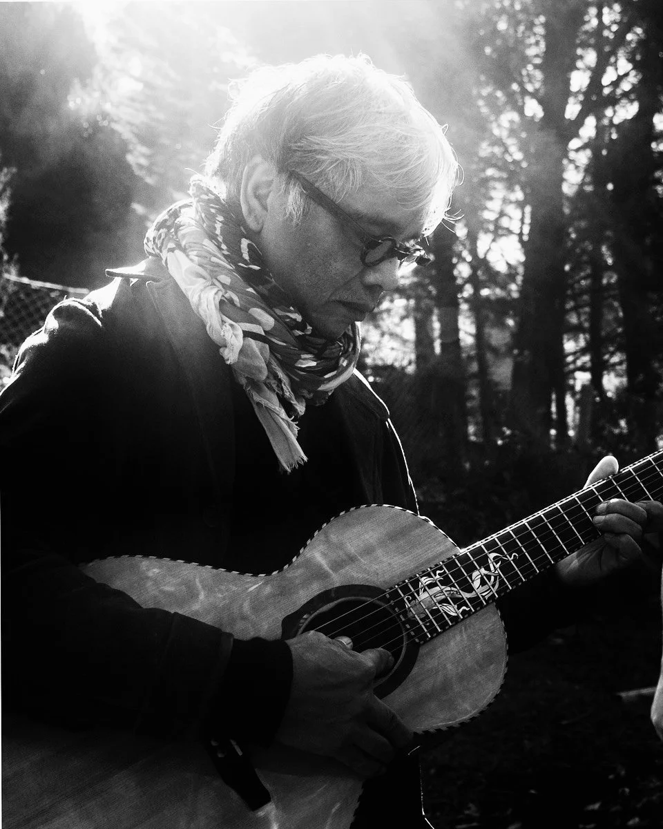 Explore TRIPLE AXEL - a revolutionary new platform designed to address taA black-and-white photo of an older man with gray hair and glasses playing an acoustic guitar outdoors, with trees in the background and sunlight filtering through the branches.