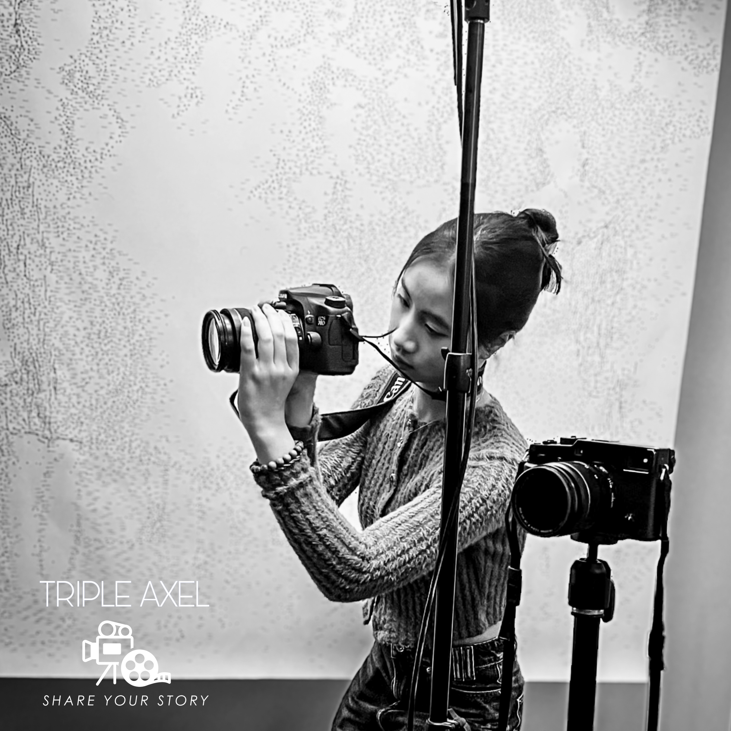 A woman with her hair in a bun takes a photograph using a professional camera mounted on a tripod. She is smiling and appears to be indoors against a textured wall with a pattern. There are graphics and text on the image that read "TRIPLE AXEL" and "SHARE YOUR STORY."