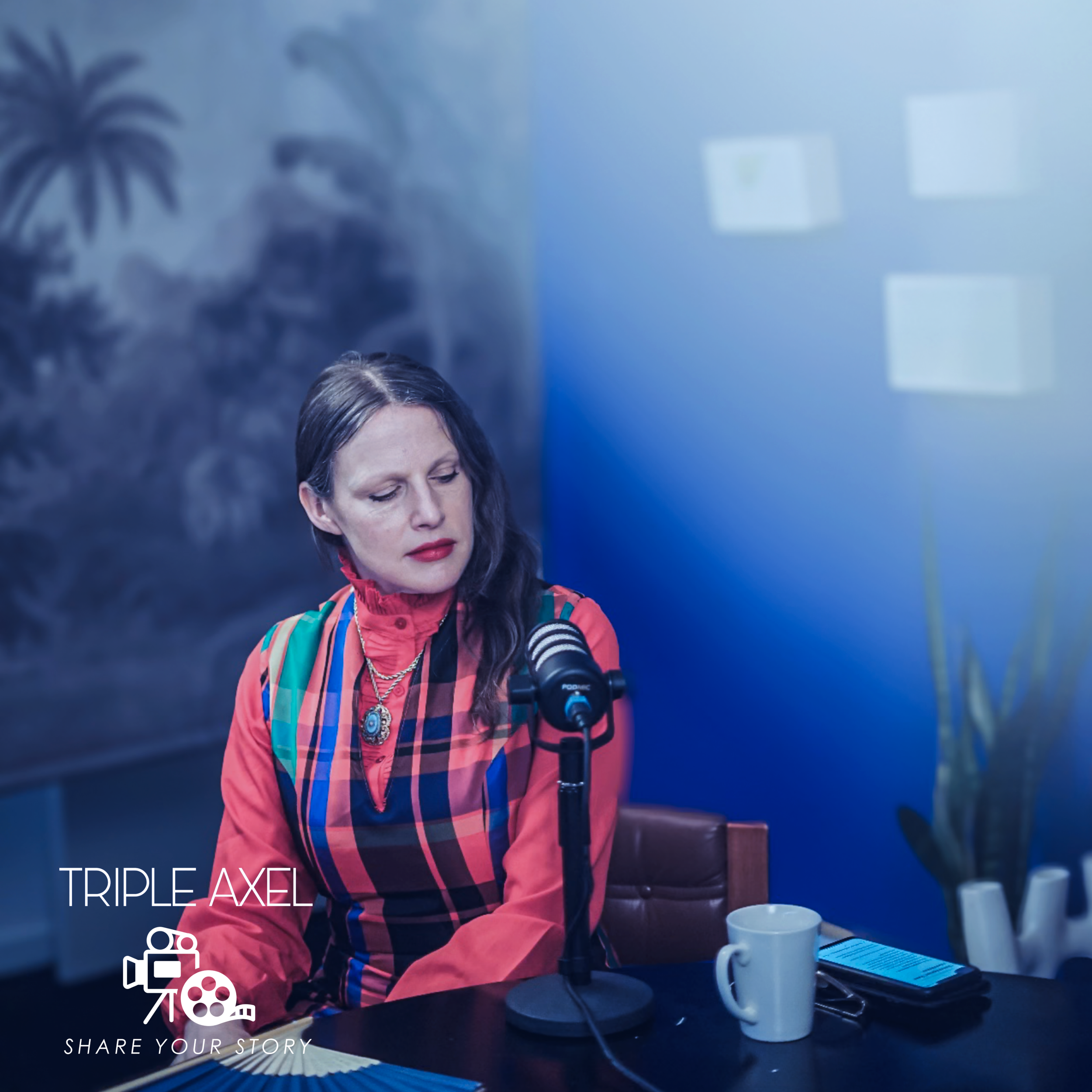 A woman with long dark hair, wearing a colorful checkered shirt and turquoise jewelry, is sitting at a table with a vintage camera and a white mug, in front of a bright blue wall with window light.