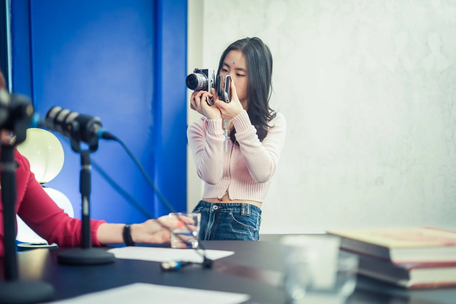 Young woman intern taking a photograph with a camera indoors, with books and a microphone on the table nearby.