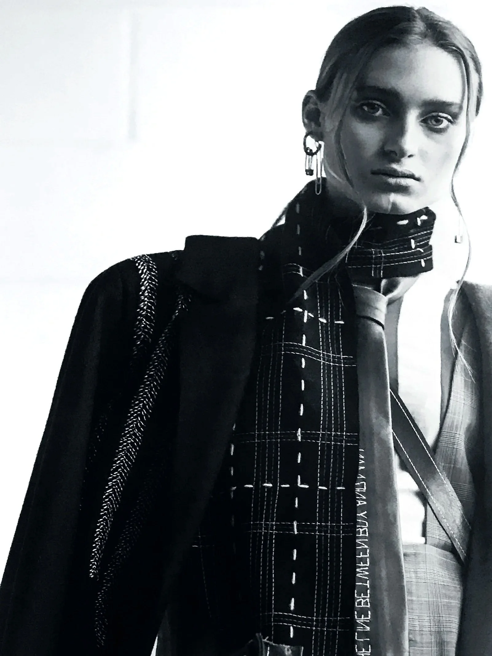 Black and white photo of a woman with styled hair wearing designer clothing, including a checkered shirt, a blazer, earrings, and a scarf, looking at the camera.