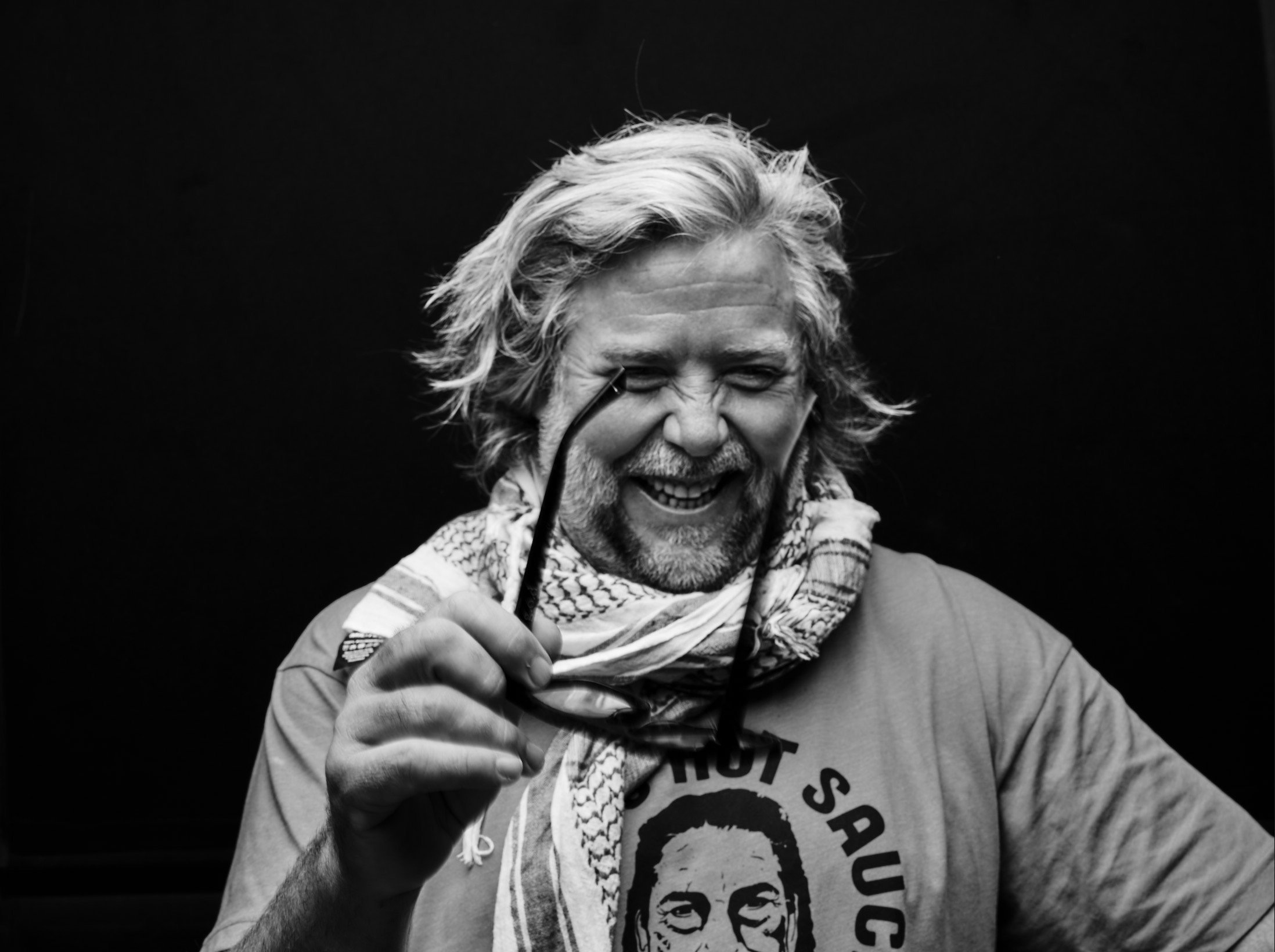 A smiling older man with long, wavy gray hair and a beard, holding a pair of glasses, wearing a graphic T-shirt and a patterned scarf, against a dark background.