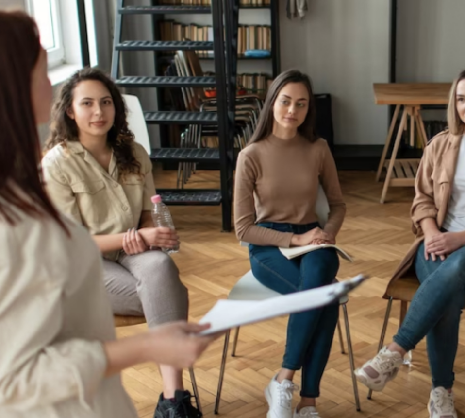 Group of women in a circle participating in a discussion or meeting in a cozy, well-lit room with bookshelves and wooden floors.