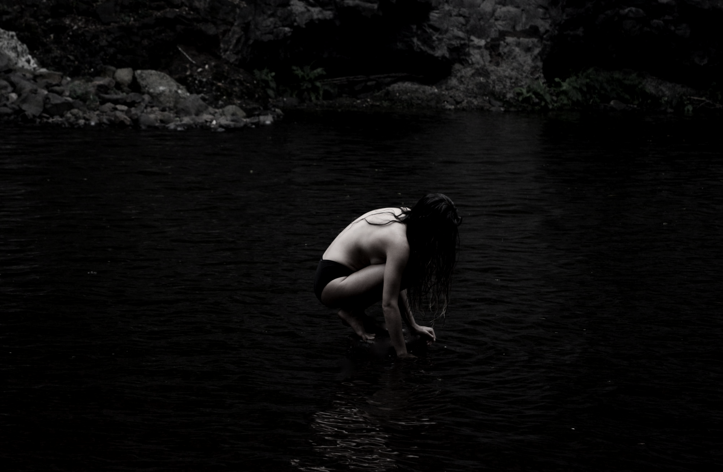 A person with long dark hair wearing black swimsuits squatting in dark water near a rocky shoreline with sparse vegetation.