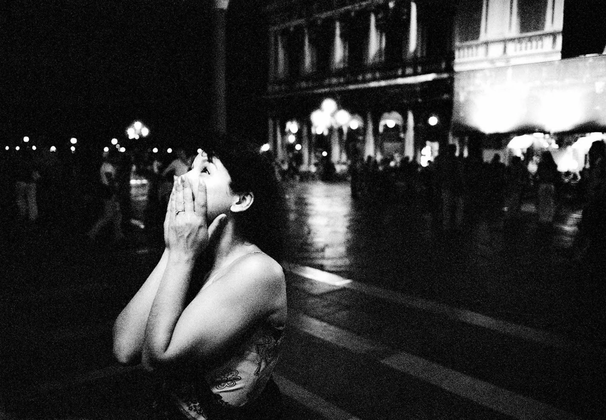 A woman with short dark hair, wearing a sleeveless top and rings, is sitting at an outdoor venue at night, covering her mouth with both hands and looking upwards, with a lively background of blurred people and illuminated buildings.