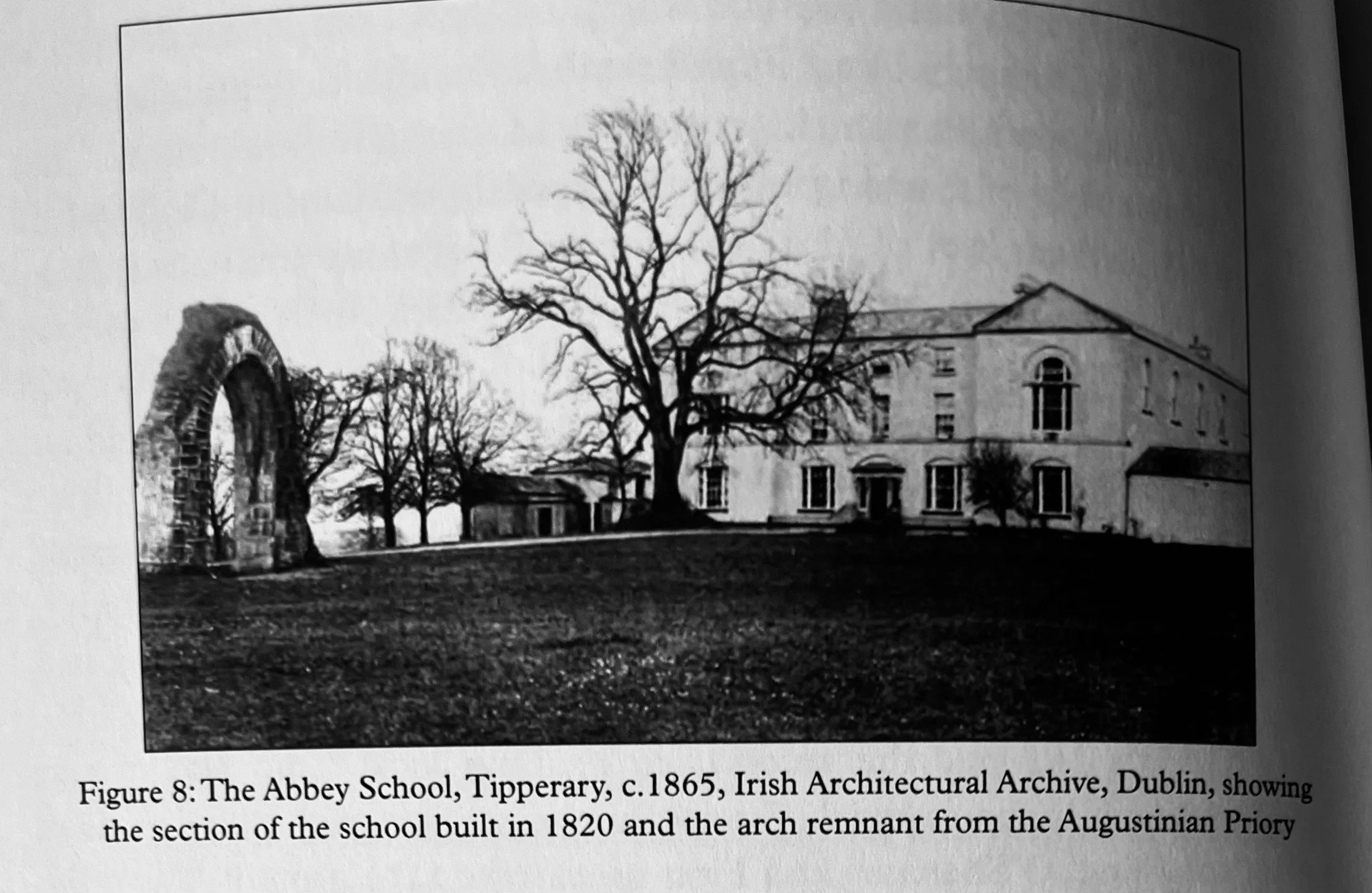 A black and white photo of a historic building called The Abbey School in Tipperary, Ireland, around 1865. The building is large with multiple windows and a pitched roof, surrounded by a large yard. A leafless tree stands prominently in the foreground. To the left, there is an arch remnant from an ancient priory.