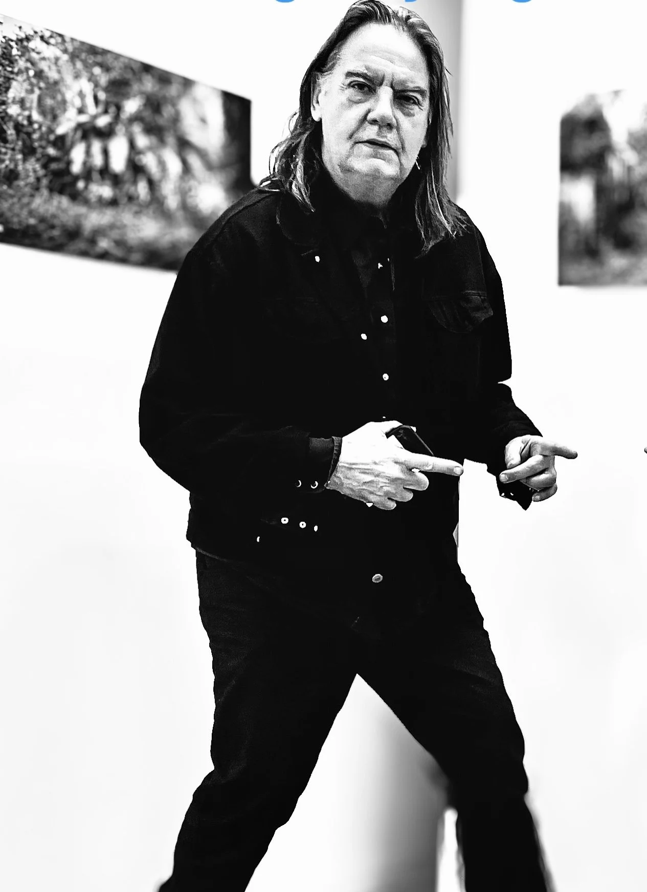 Explore TRIPLE AXEL - a revolutionary new platforBlack and white photo of a person with long hair, wearing a dark jacket, standing in front of a white wall with framed photographs. The person is pointing with both hands, and has a serious expression.