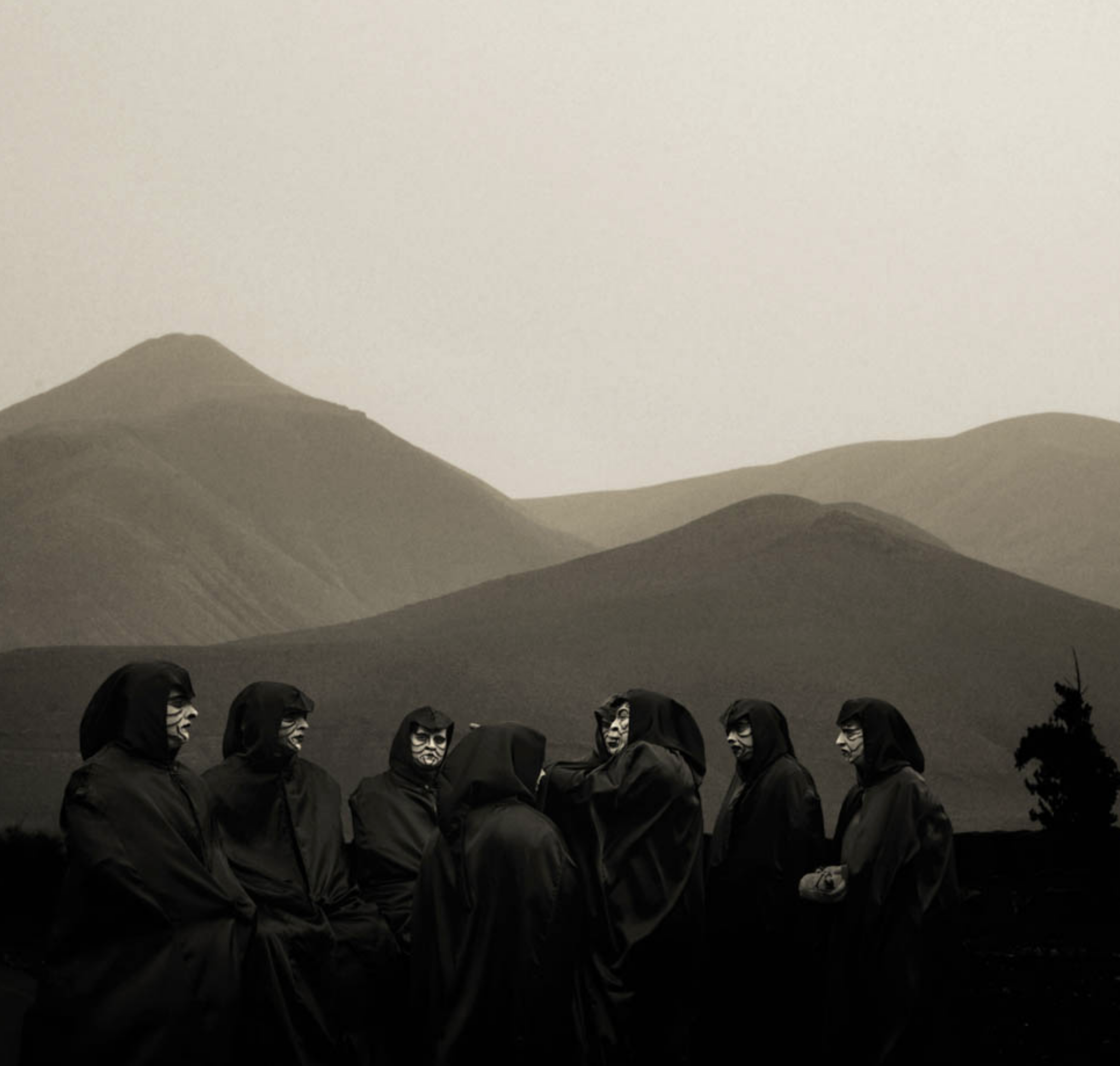 A group of nine people wearing dark hooded cloaks and masks standing in a line outdoors with rolling hills in the background.